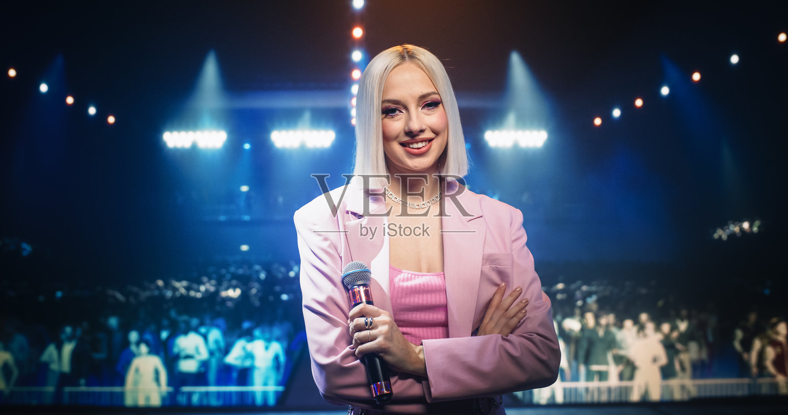 美丽女艺术家在音乐会上站在舞台上，摆姿势并看向镜头。年轻的励志歌手双臂交叉，微笑着手持麦克风。照片摄影图片