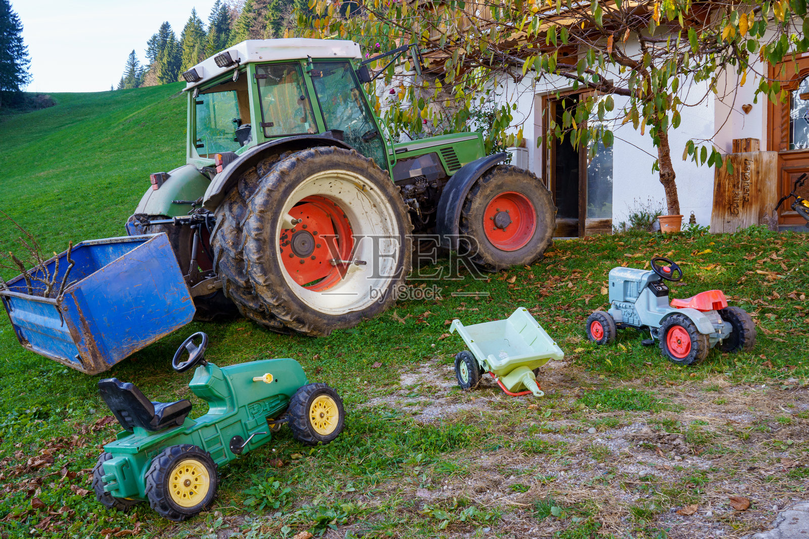 两个儿童脚踏拖拉机和一个拖车停在一辆红轮绿色拖拉机和拖车前面。背景是一座农舍和草地，呈现出丘陵的风景。照片摄影图片