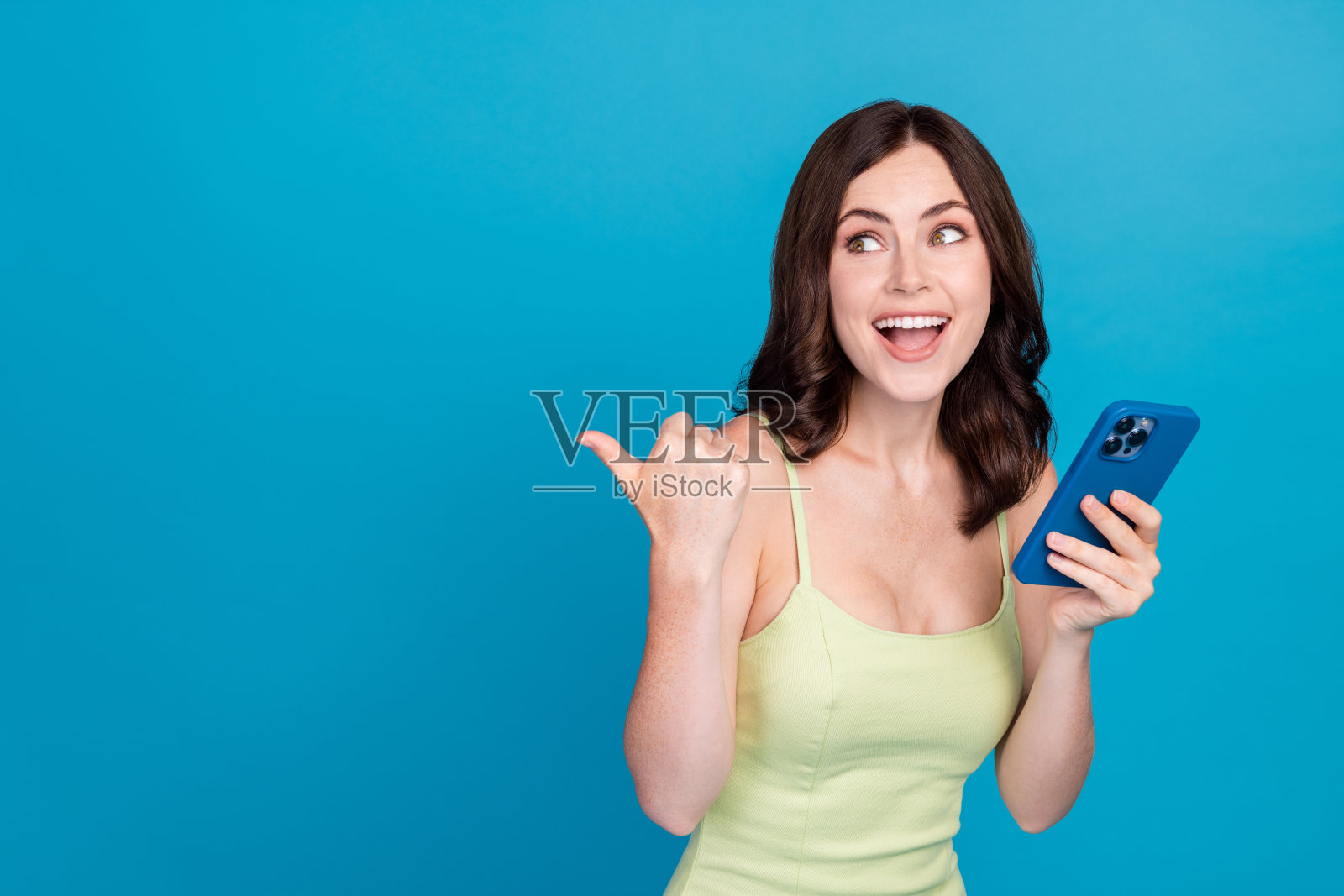 穿着绿色服装的美丽梦幻女士，正在与现代设备聊天，指着空白空间，背景为蓝色。照片摄影图片