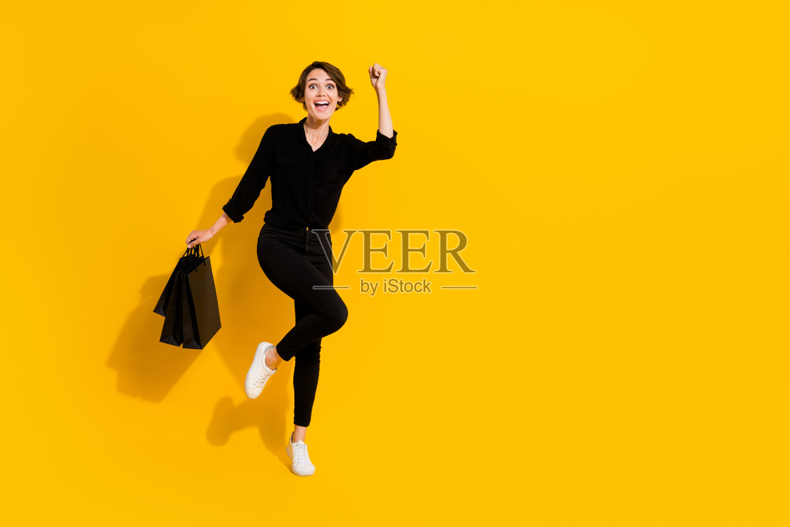 惊讶的年轻女士全身照，手持购物袋，穿着衬衫、牛仔裤和靴子，背景为黄色。照片摄影图片