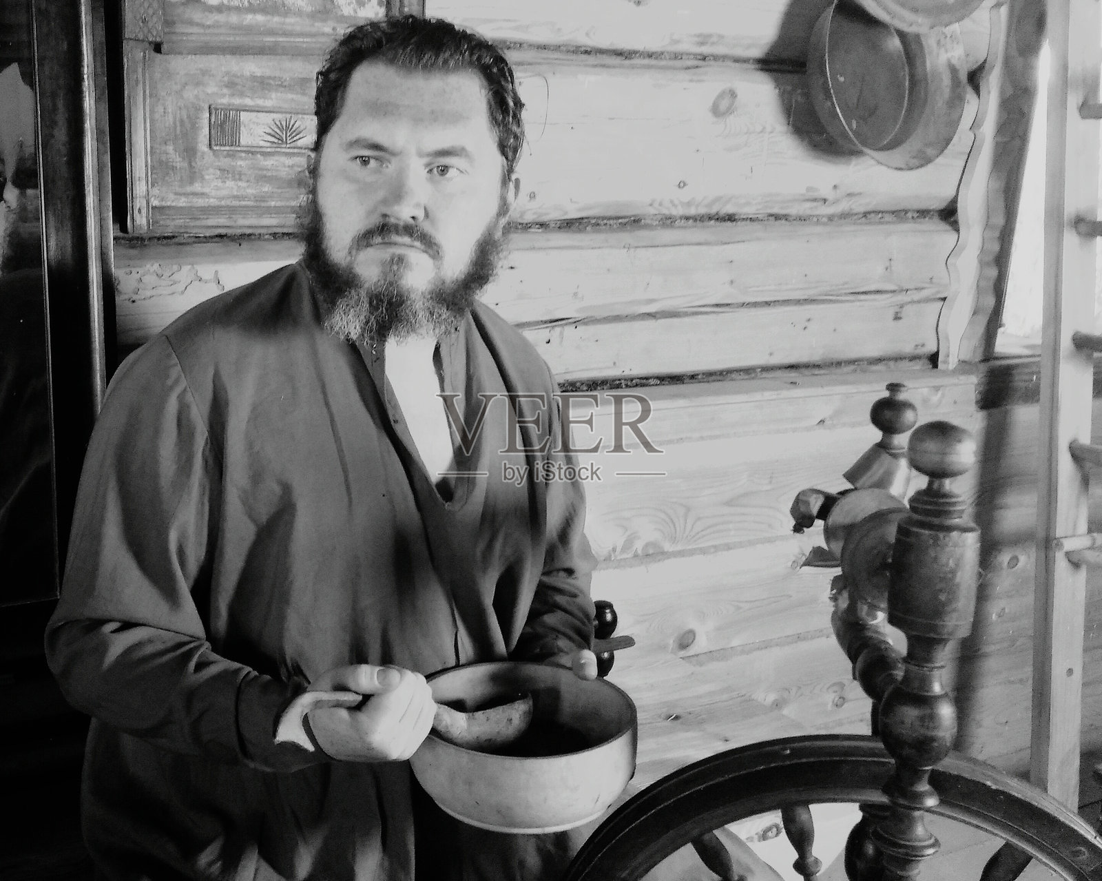 男人在家中靠着木墙，手里拿着容器，目光投向一旁。照片摄影图片