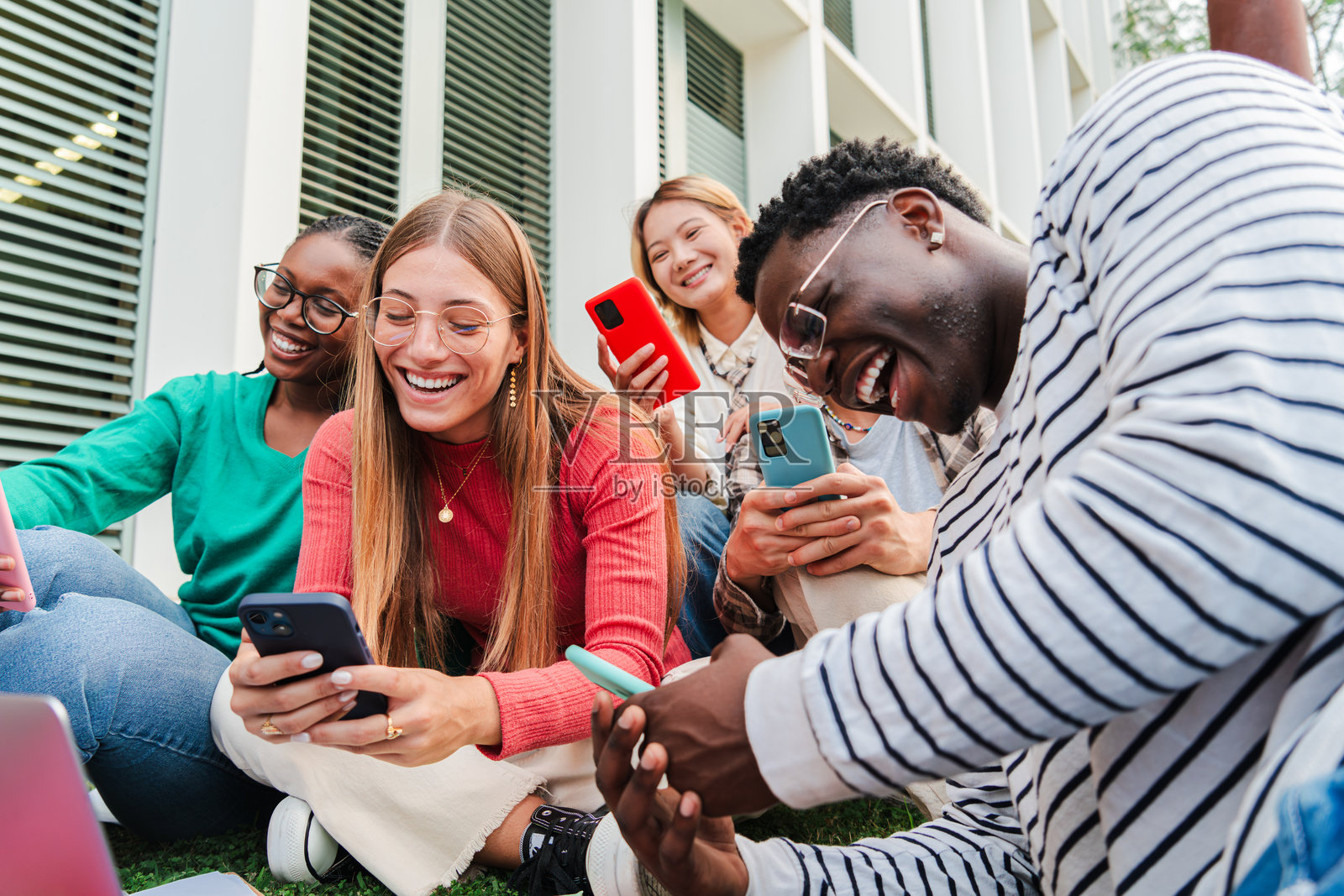 一群微笑的年轻多民族高中生正在使用手机。男生和女生们在大学校园的草坪上坐在一起，享受着社交媒体。照片摄影图片