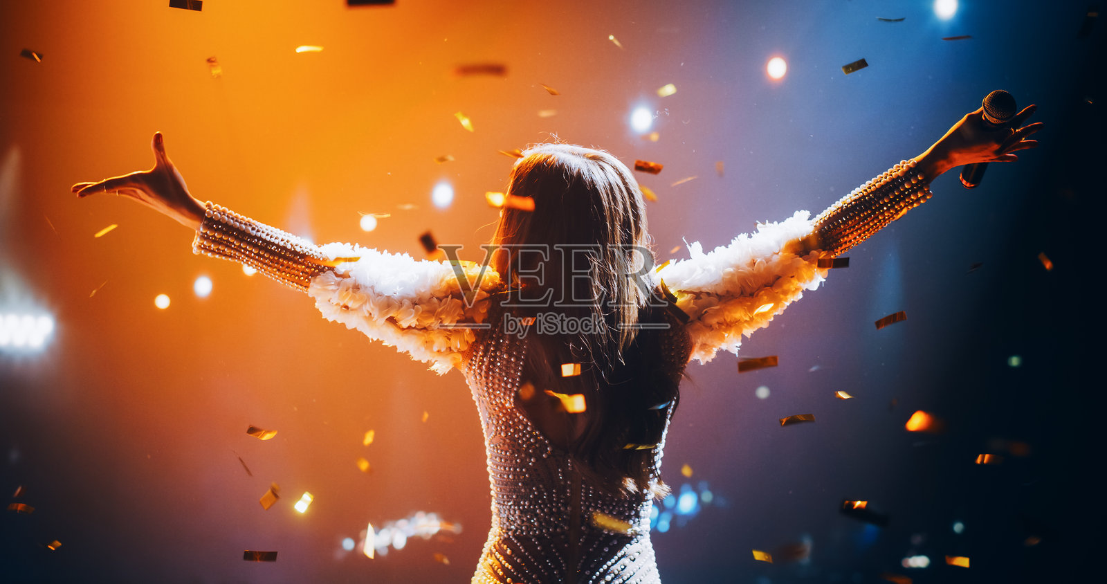 女歌手身穿引人注目的银色服装演出。才华横溢的流行歌星的壮观音乐会。史诗般的电影慢动作镜头，五彩缤纷的彩纸在舞台上的艺术家身上飘落。照片摄影图片