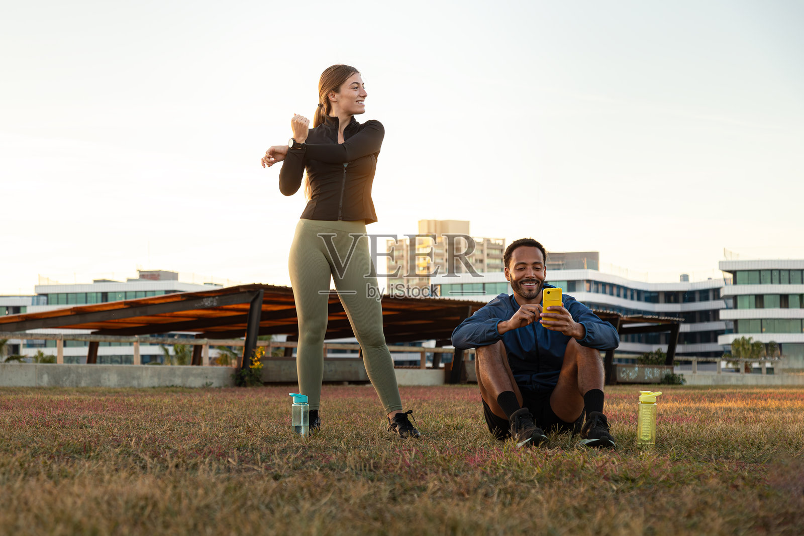 年轻的多种族情侣在城市公园跑步和锻炼后，伸展身体并查看手机。照片摄影图片