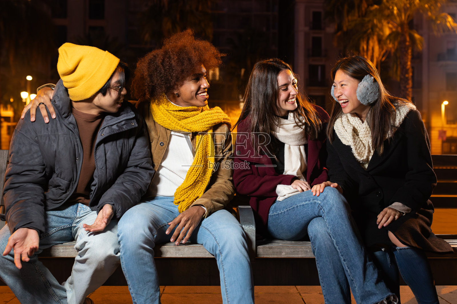 一群多种族的朋友在寒冷的冬夜坐在城市长椅上，欢笑聊天，享受快乐时光。照片摄影图片