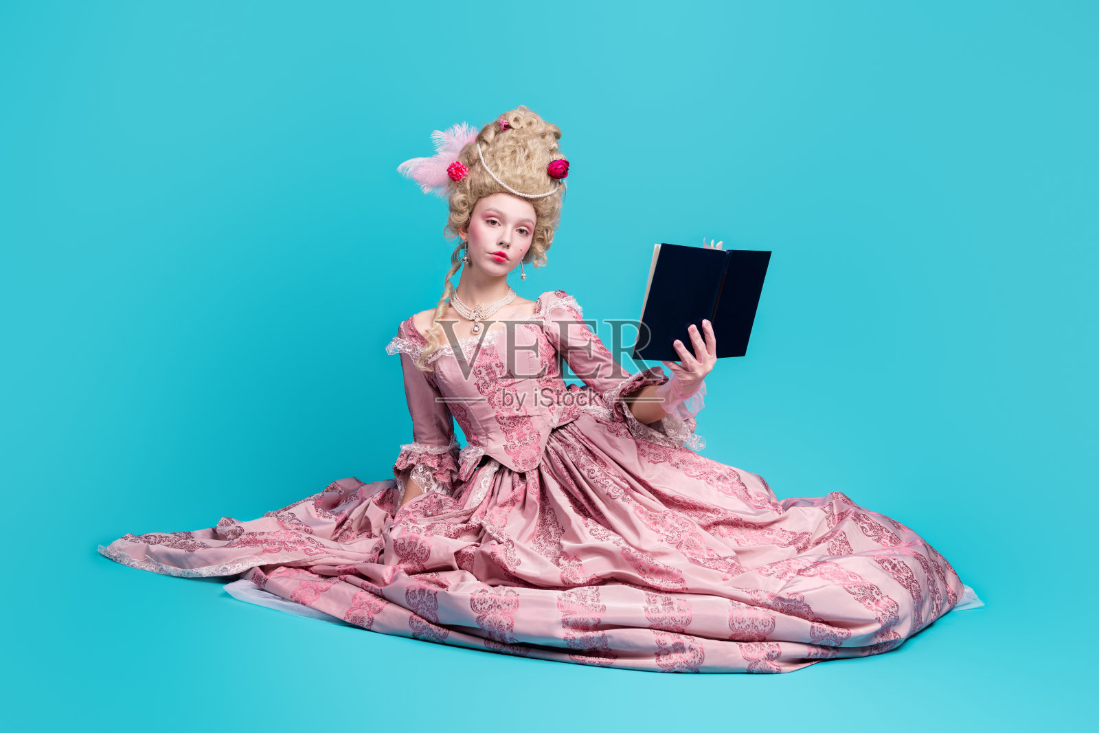 美丽迷人的公主女王穿着华丽的裙子坐在地上阅读书籍，背景为蓝色。照片摄影图片