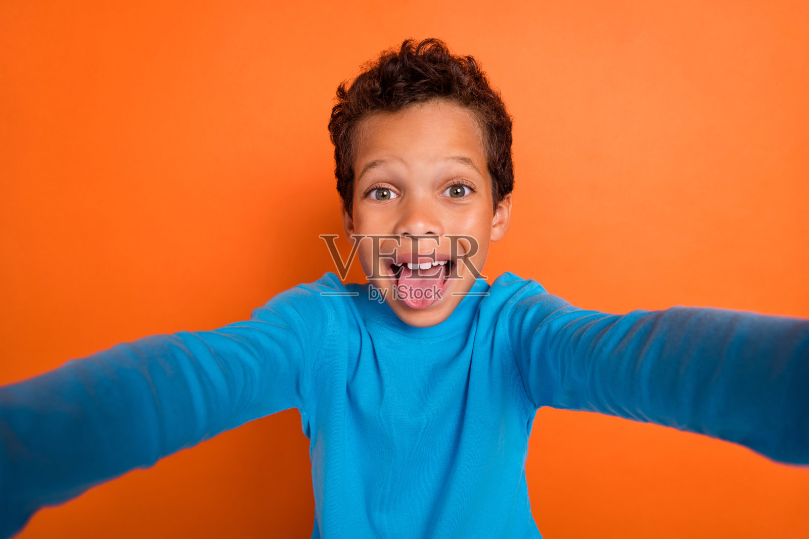 迷人的小男孩自拍吐舌头，穿着时尚的蓝色服装，表现出不礼貌的行为，背景为橙色。照片摄影图片