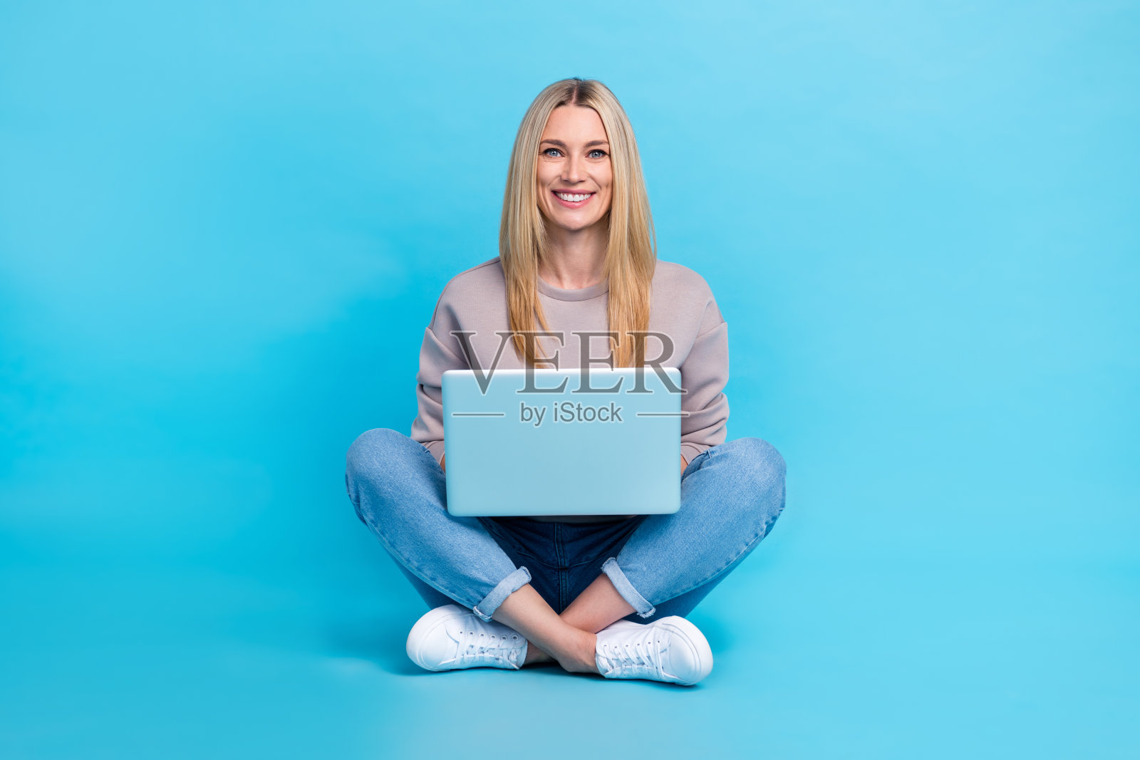 穿着灰色卫衣的漂亮甜美女士全身照，正在使用现代电子设备，背景为蓝色。照片摄影图片