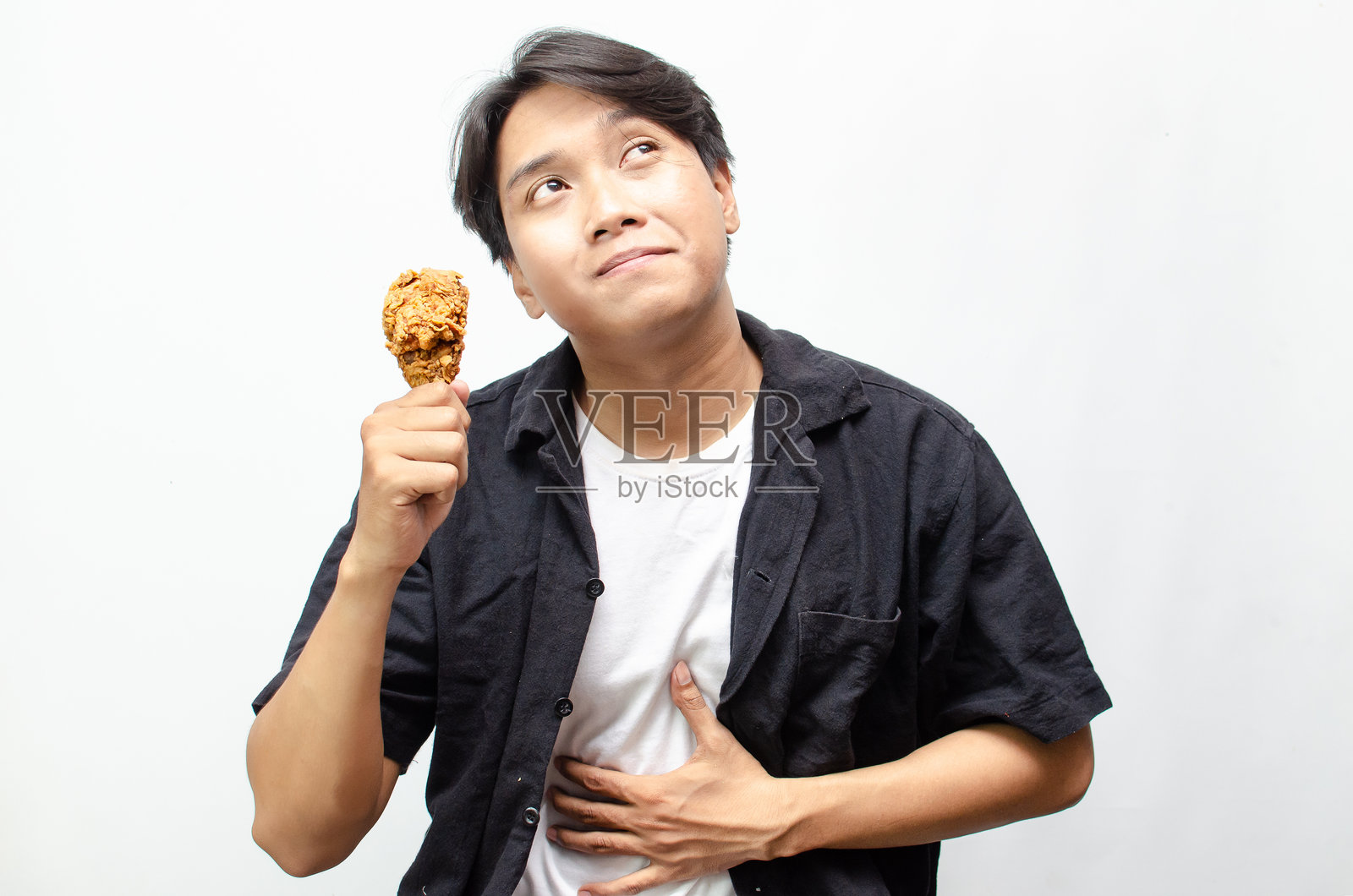 饥饿的年轻亚洲男子正在享用香脆炸鸡腿，脸上露出满意的表情。照片摄影图片