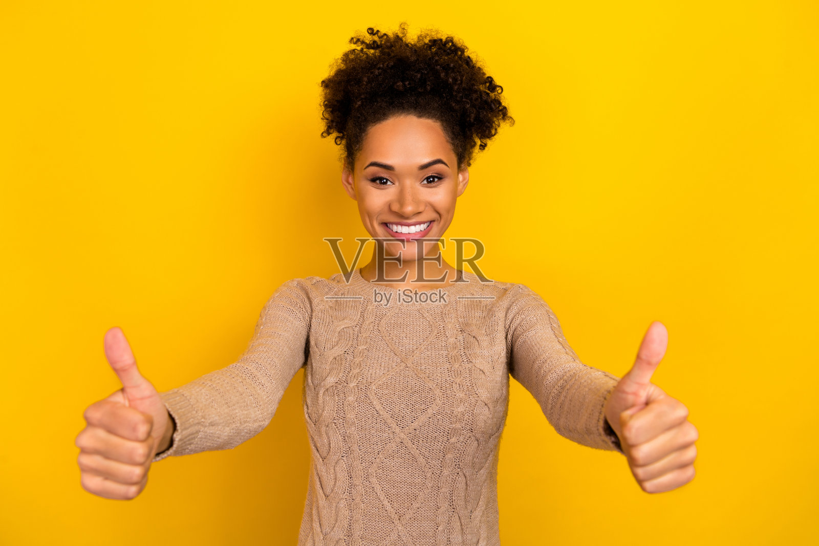 可爱的年轻棕发女孩竖起大拇指，穿着米色毛衣，背景为黄色。照片摄影图片