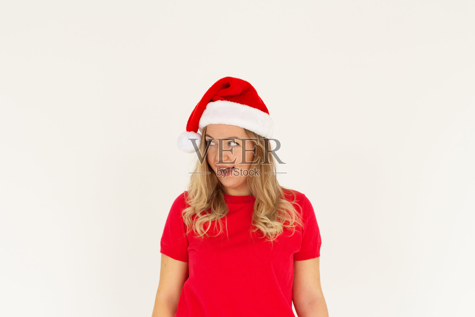 惊讶的年轻圣诞老人女性，戴着圣诞帽，微笑着侧目，背景为白色，工作室肖像。快乐的新年庆祝，愉快的节日概念。照片摄影图片