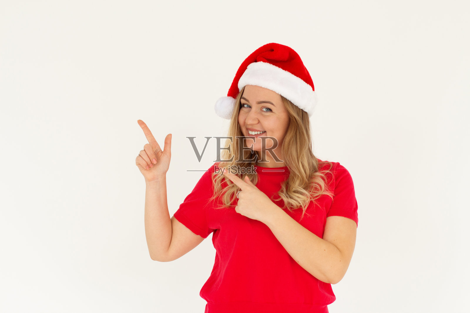 微笑的年轻圣诞女郎穿着T恤和圣诞帽，手指指向旁边的空白区域，背景为白色，工作室肖像。快乐的新年假期概念照片摄影图片