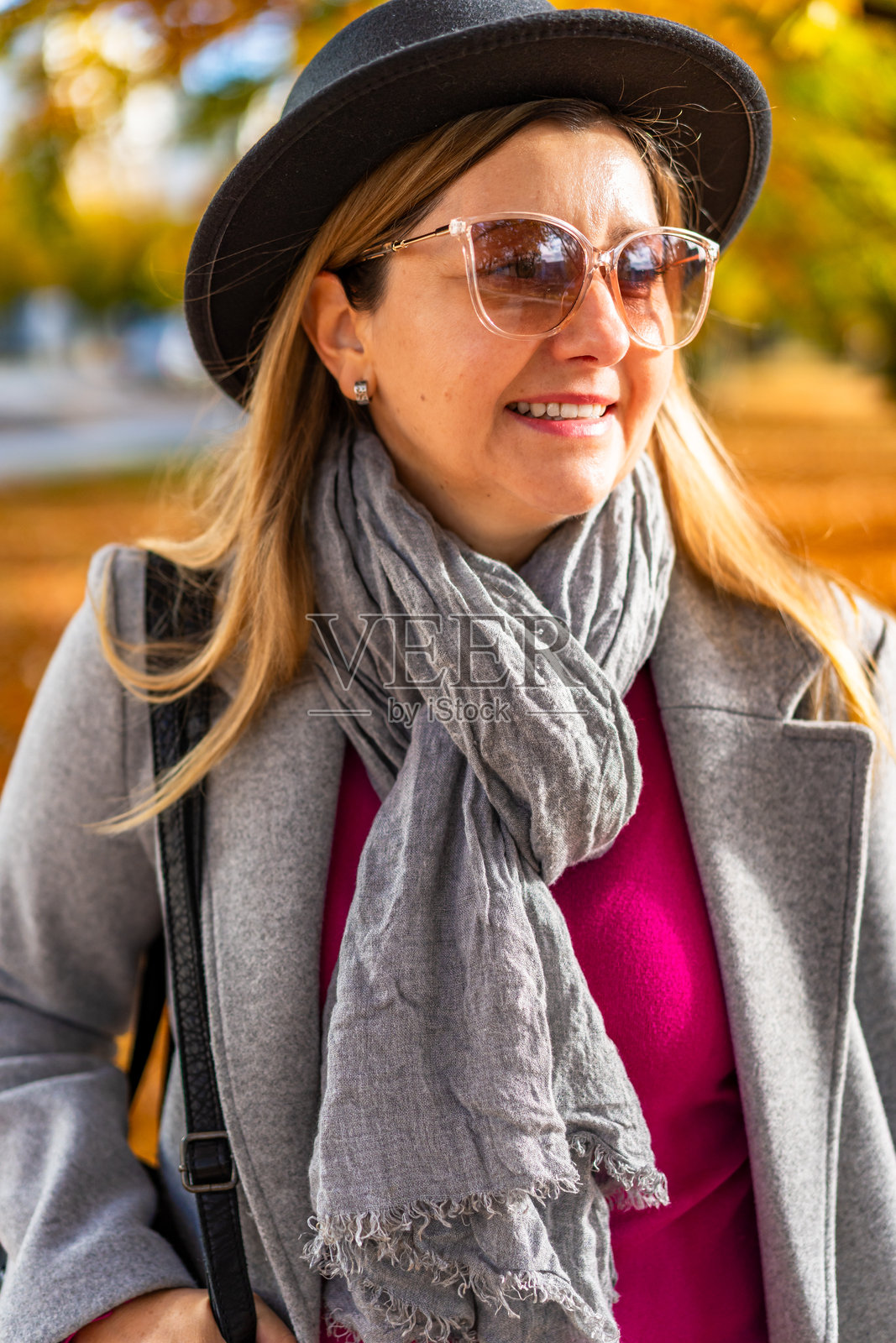美丽的年轻女性穿着牛仔裤、灰色外套、格子温暖围巾、黑色边帽和太阳镜，在阳光明媚的秋日里沿着城市公园的小巷走，周围是落叶树。正面视角。照片摄影图片