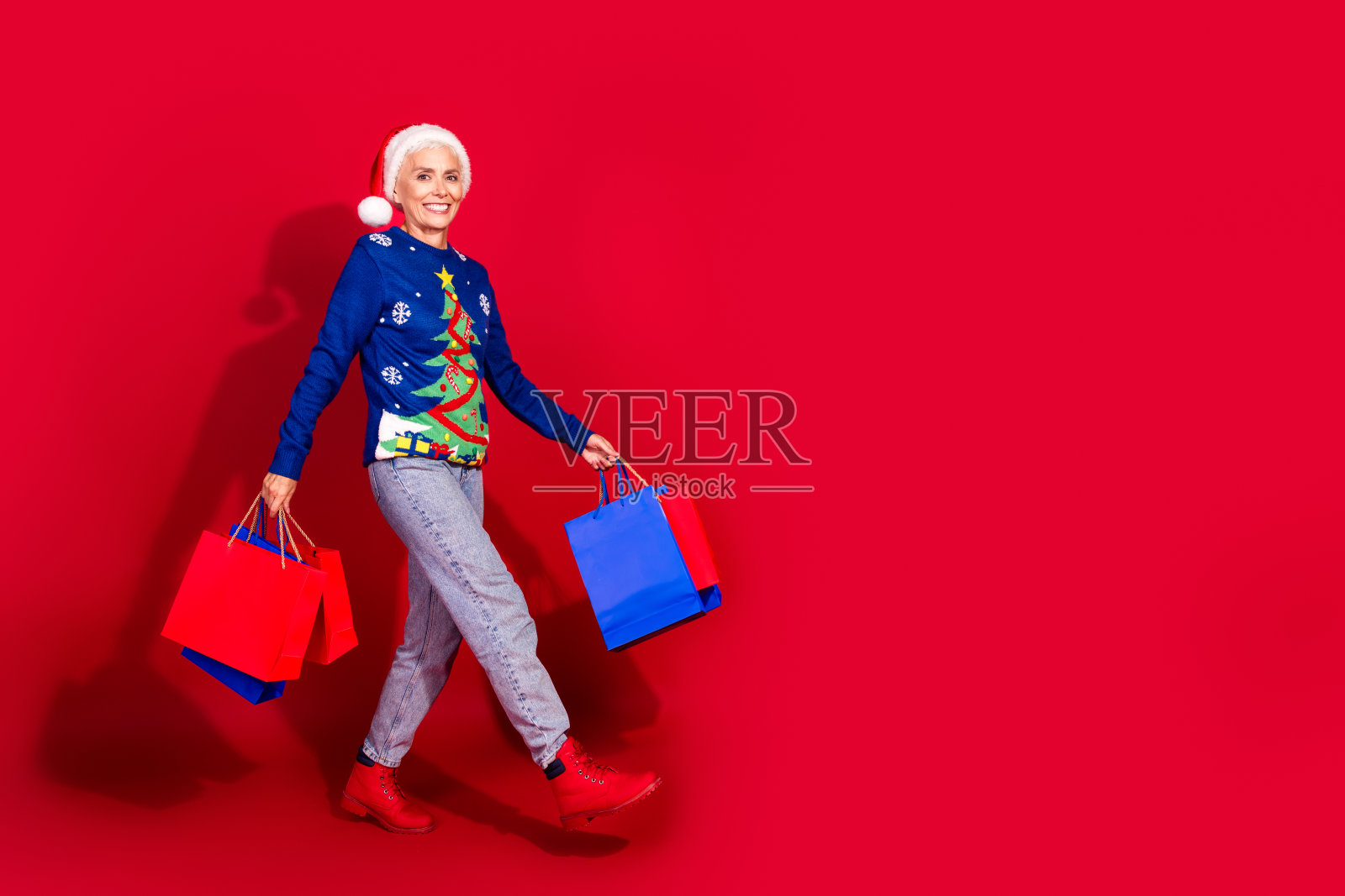 全身肖像的美丽老年女士手持购物袋，走在空旷的空间中，背景为鲜艳的红色，迎接新年和圣诞节。照片摄影图片