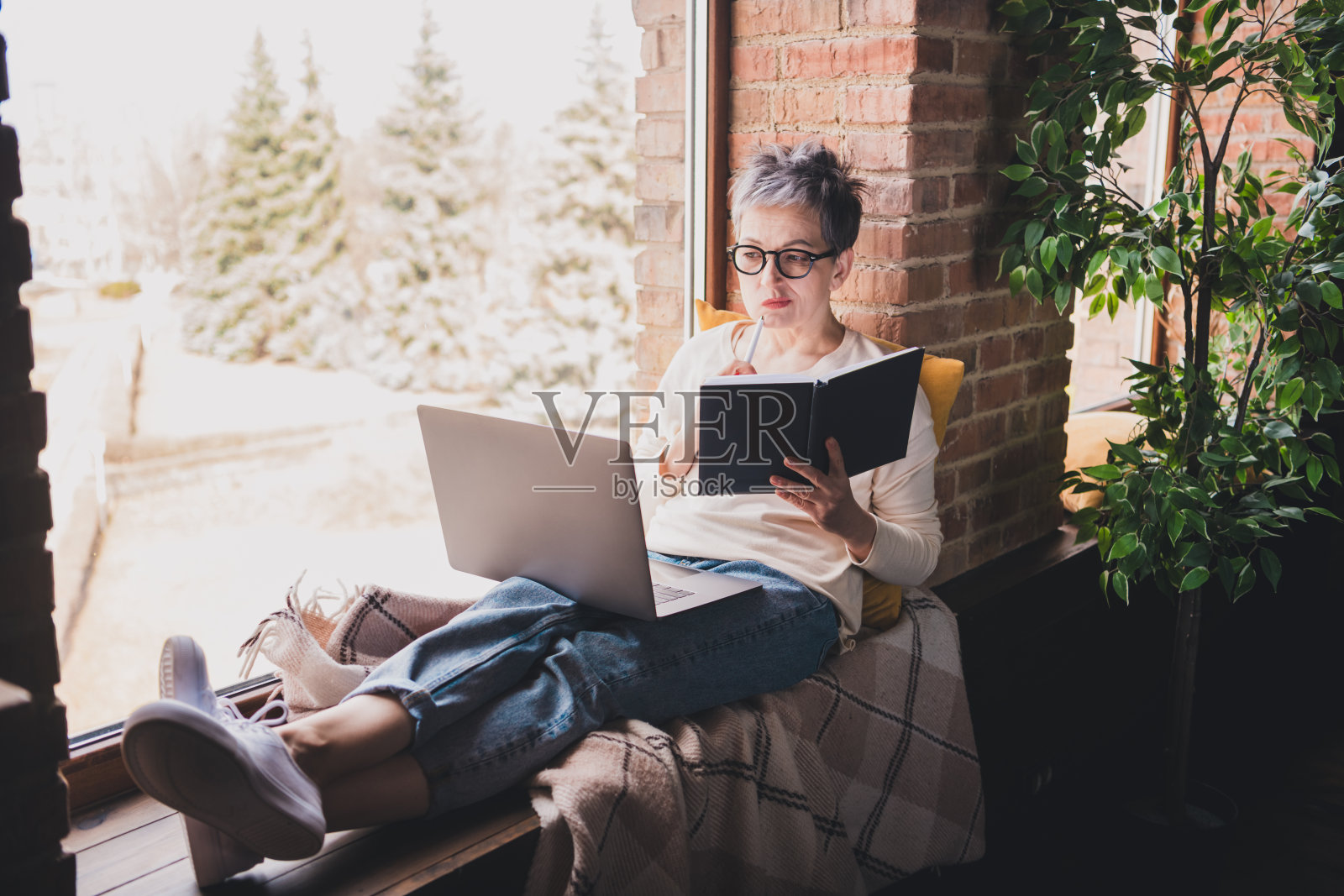 女性，短灰发，穿着休闲风格，坐在室内，享受着与笔记本电脑和笔记本一起的闲暇时光，窗边。照片摄影图片