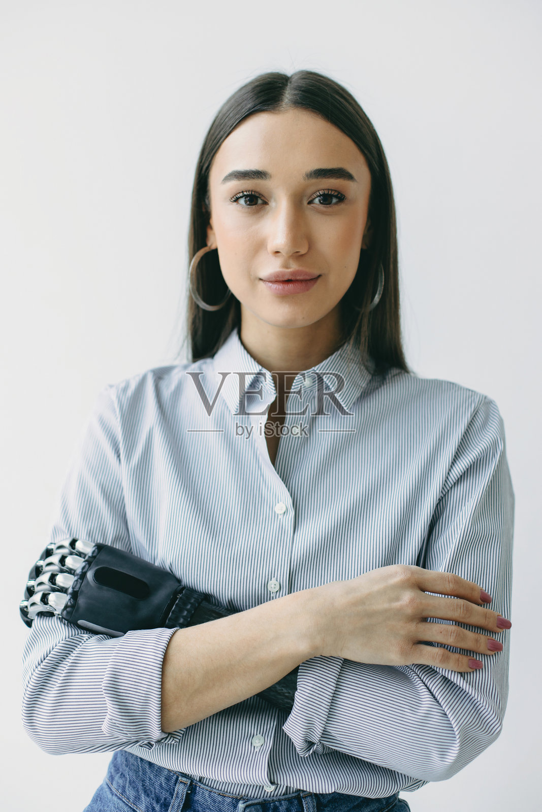 时尚年轻女性穿着衬衫和牛仔裤，双臂交叉，处于封闭姿势，手上有3D打印的赛博仿生手，面带平静积极的表情看向镜头。照片摄影图片