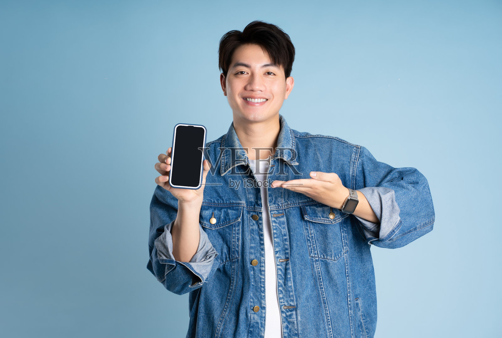 一位亚洲男士穿着夹克，手持手机在蓝色背景前摆姿势的肖像。照片摄影图片