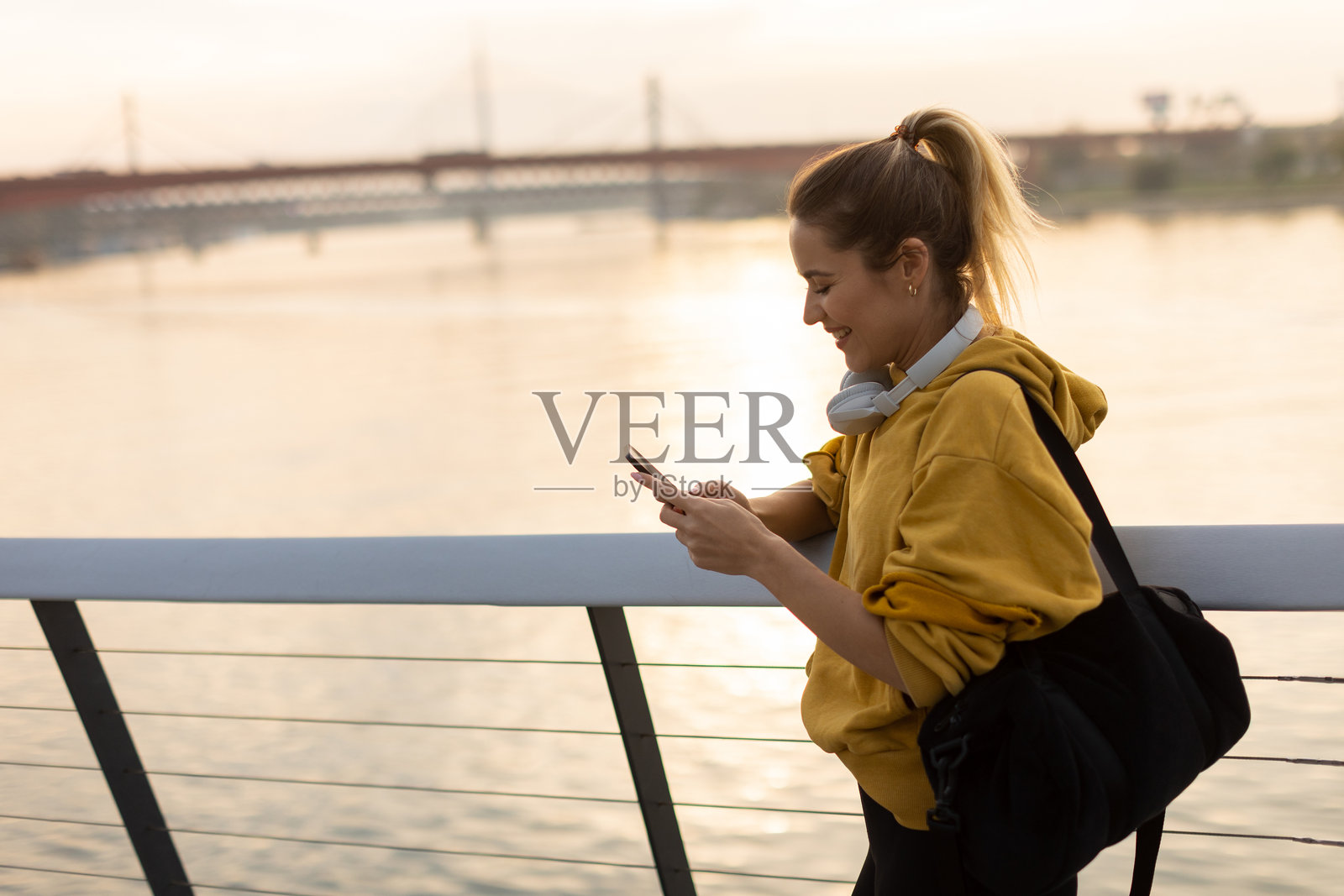 一位穿着黄色连帽衫的女性在宁静的城市环境中，享受着日落时分在河边查看手机的宁静时刻。照片摄影图片