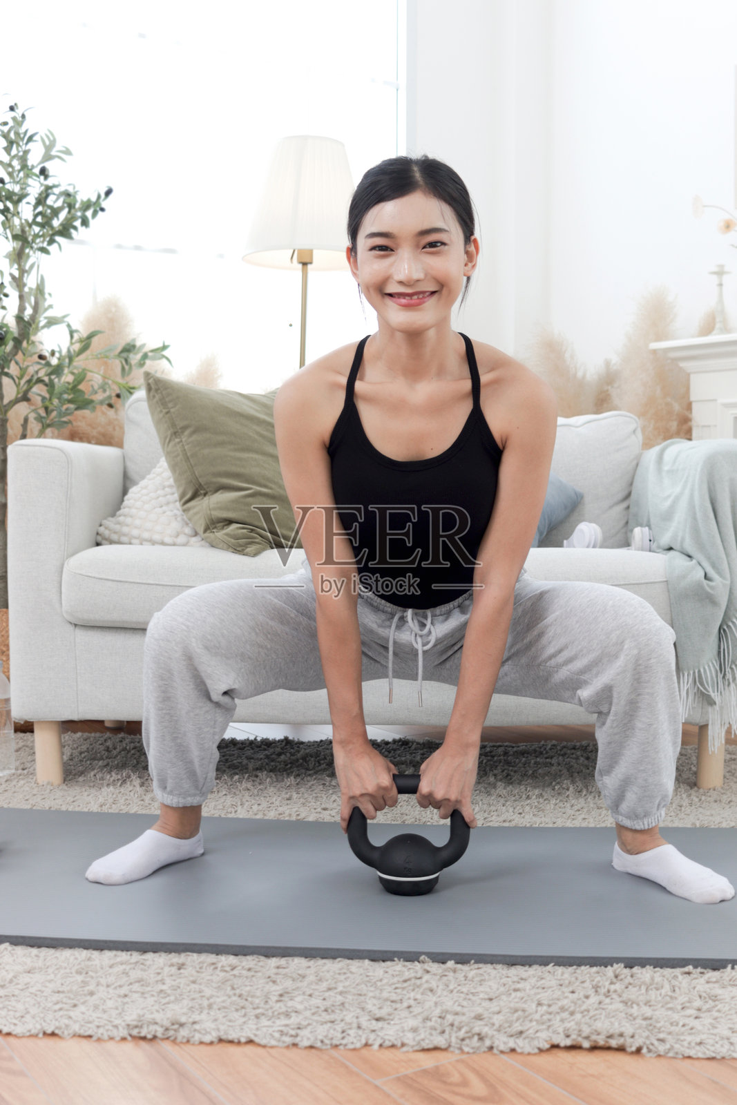 亚洲美丽女性在客厅的瑜伽垫上使用壶铃锻炼。快乐的女性在家举起壶铃进行锻炼。运动女孩在进行体育锻炼。健康与运动爱好。照片摄影图片