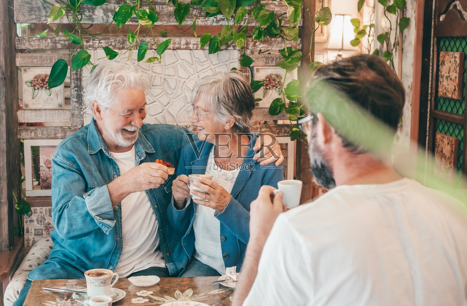 快乐的老年夫妇和成熟的儿子在舒适的咖啡馆里休息，享用咖啡、卡布奇诺和水果蛋糕。三个人一起享受美好时光。照片摄影图片