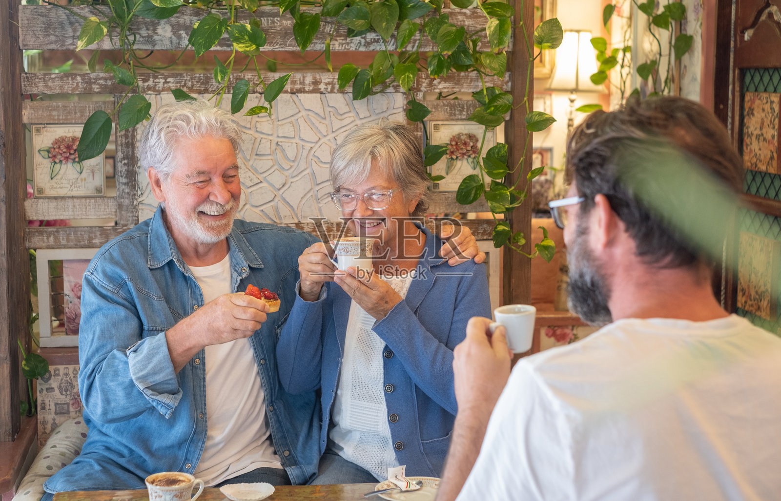 快乐的老年夫妇和成熟的儿子在舒适的咖啡馆里休息，享用咖啡、卡布奇诺和水果蛋糕。三个人一起享受美好时光。照片摄影图片