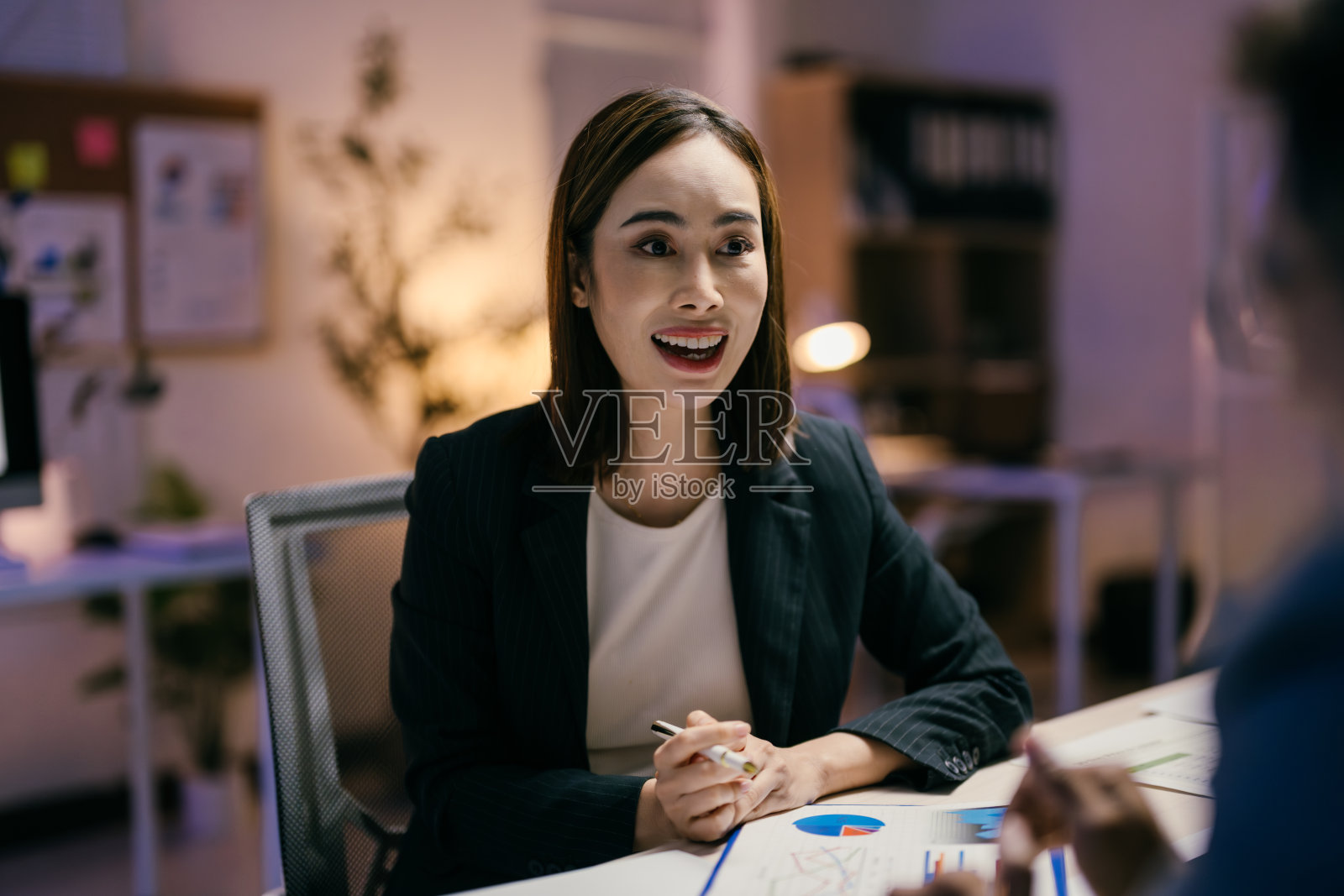 年轻女商人在办公室的桌子前坐着，手握一支笔，与同事讨论图表。照片摄影图片