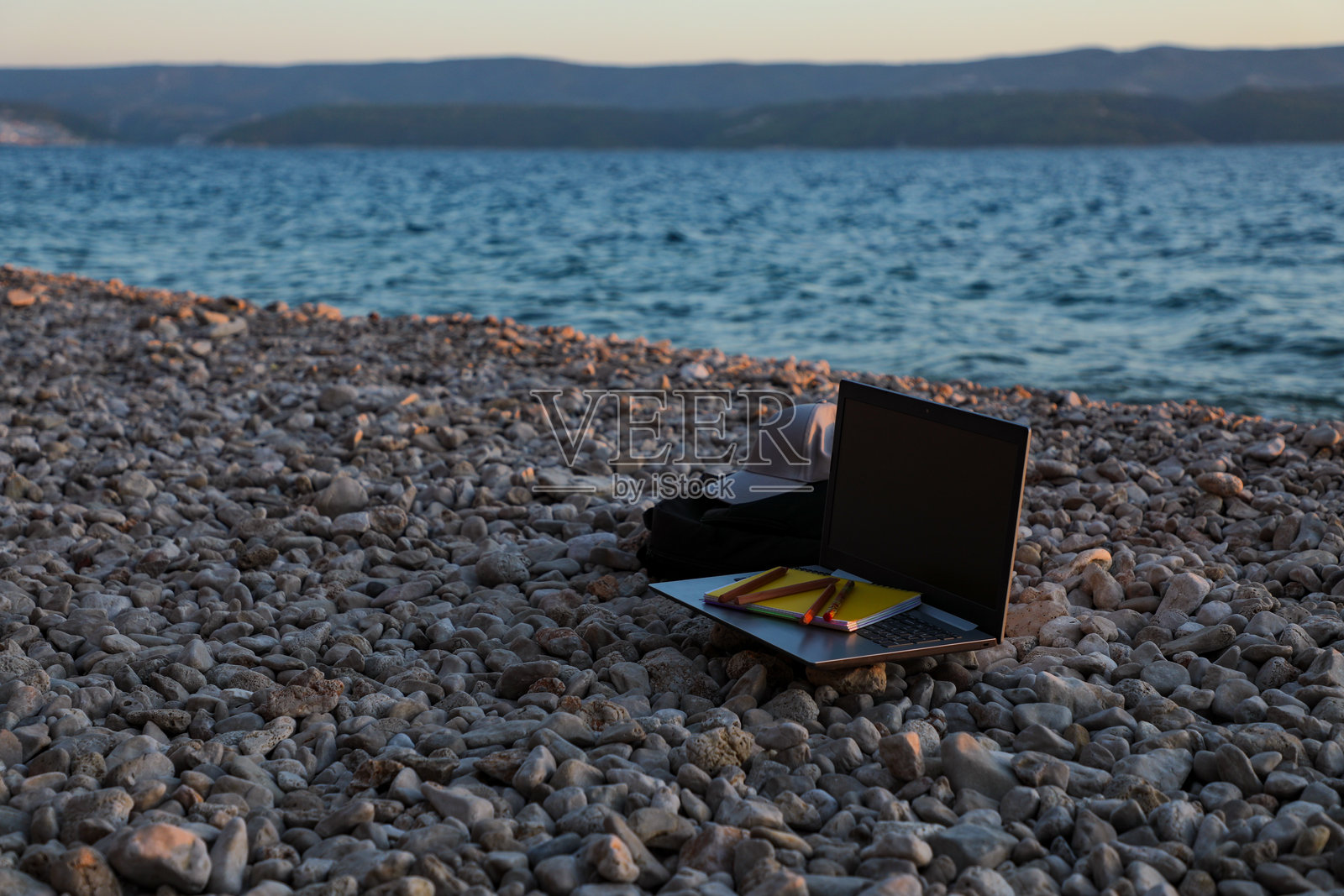 在海边的卵石沙滩上，日落时分有一台笔记本电脑、一顶棒球帽、一只笔记本包、一份笔记本和几支铅笔。照片摄影图片