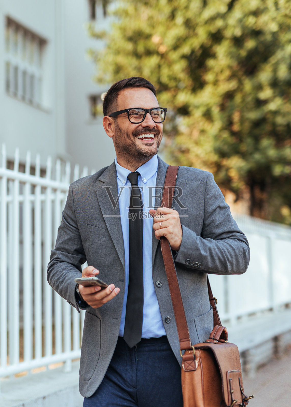 自信的商人在户外走动，手里拿着智能手机和包。照片摄影图片