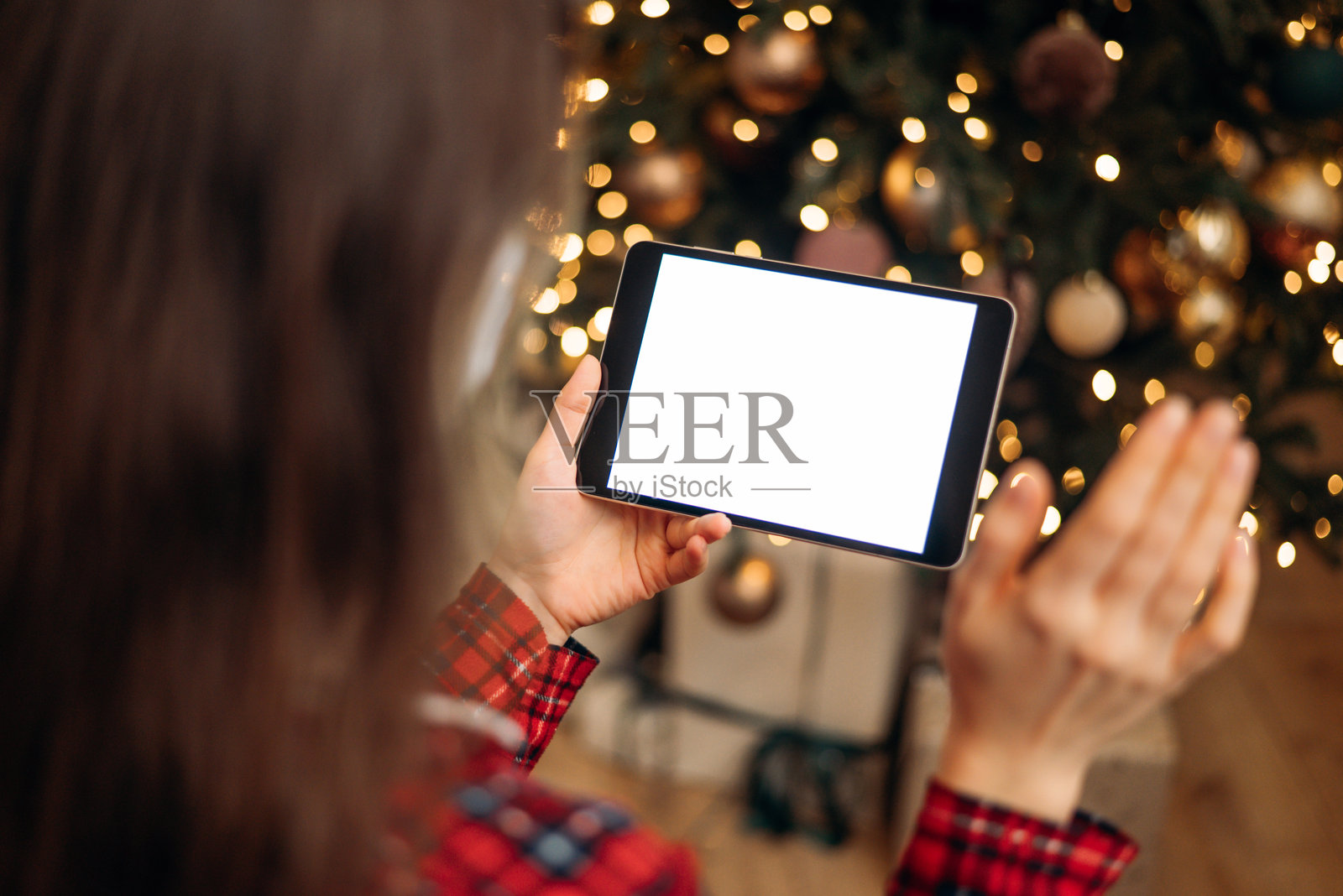 女人在圣诞树前手持空白屏幕的平板电脑，背景中有节日的散景灯光，准备进行假日视频通话或虚拟问候。照片摄影图片