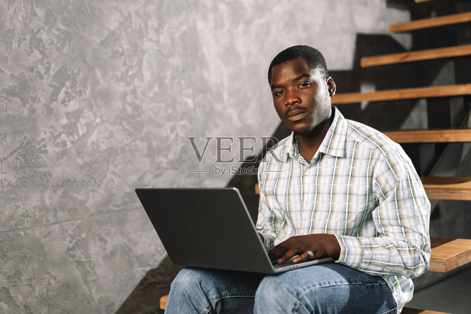 一个非洲年轻人坐在木梯上，在房间里拿着笔记本电脑照片摄影图片