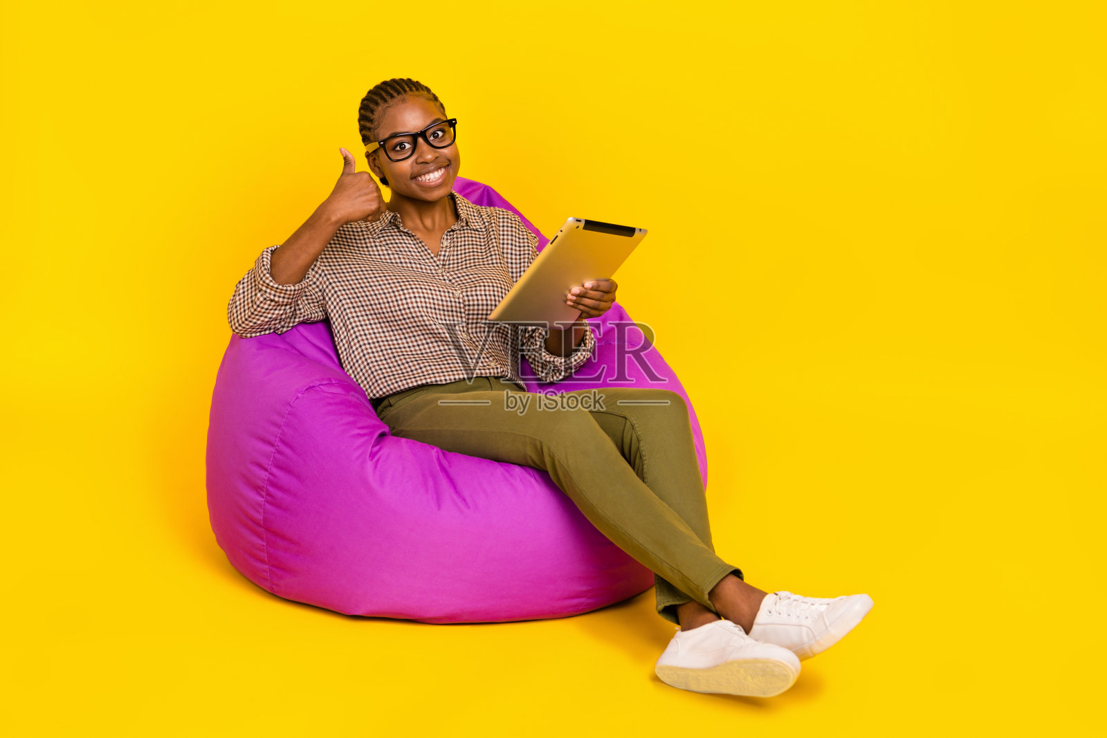 自信漂亮的女性穿着格子衬衫，坐在豆袋椅上，竖起大拇指，手持现代设备，背景为空白的黄色。照片摄影图片
