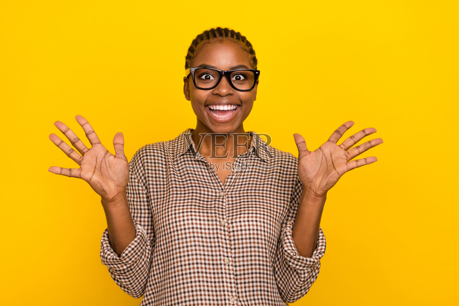 可爱的惊讶女性穿着格子衬衫，戴着眼镜，微笑着举起双臂，背景为黄色。照片摄影图片