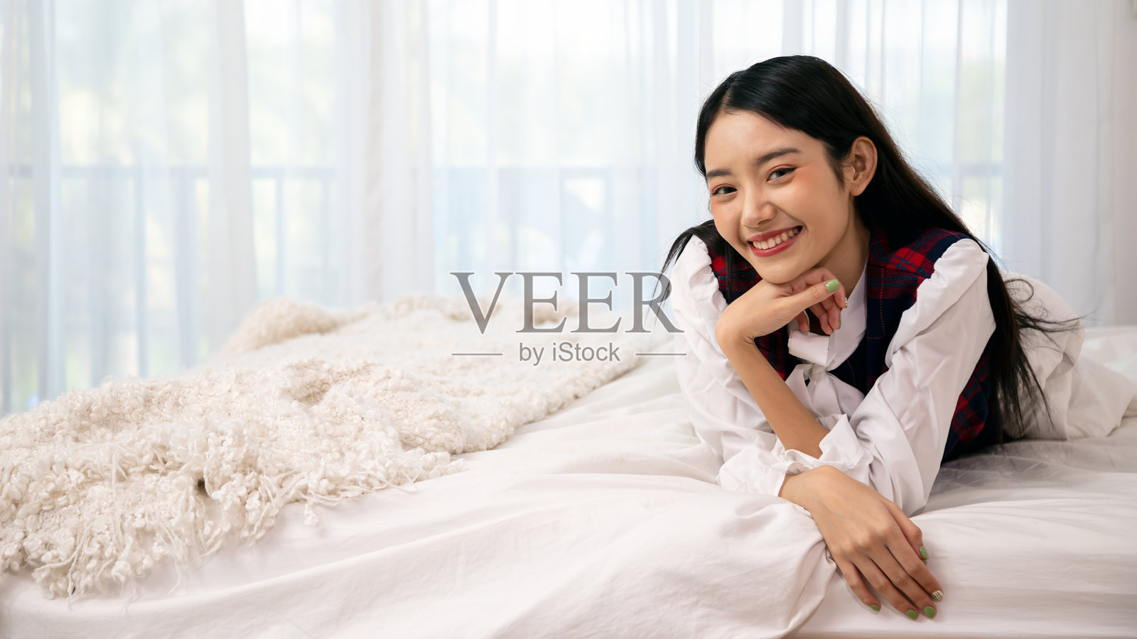 年轻的亚洲女性微笑着躺在舒适的床上，身上盖着蓬松的白色毯子，享受着宁静愉快的时光，留有空白空间。照片摄影图片