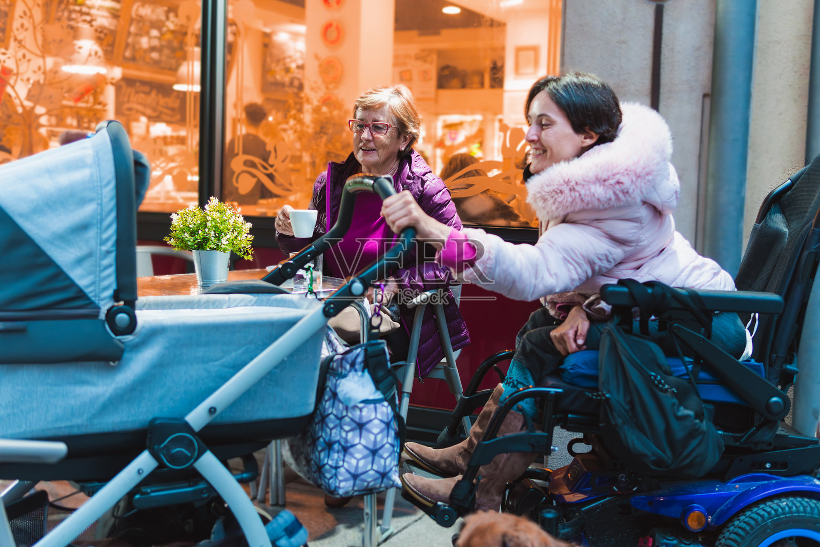 母亲和女儿坐在咖啡馆露台上，开心而温柔地看着婴儿车里的宝宝，享受着咖啡。概念：残障人士的生活方式照片摄影图片