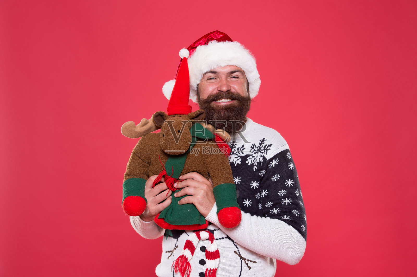 圣诞准备概念。时髦的胡子男士穿着冬季毛衣。善良与慈善。冬季假期。购买礼物。冬季促销。冬季盛宴。驯鹿玩具。戴圣诞老人帽的男人手持圣诞礼物，背景为红色。照片摄影图片