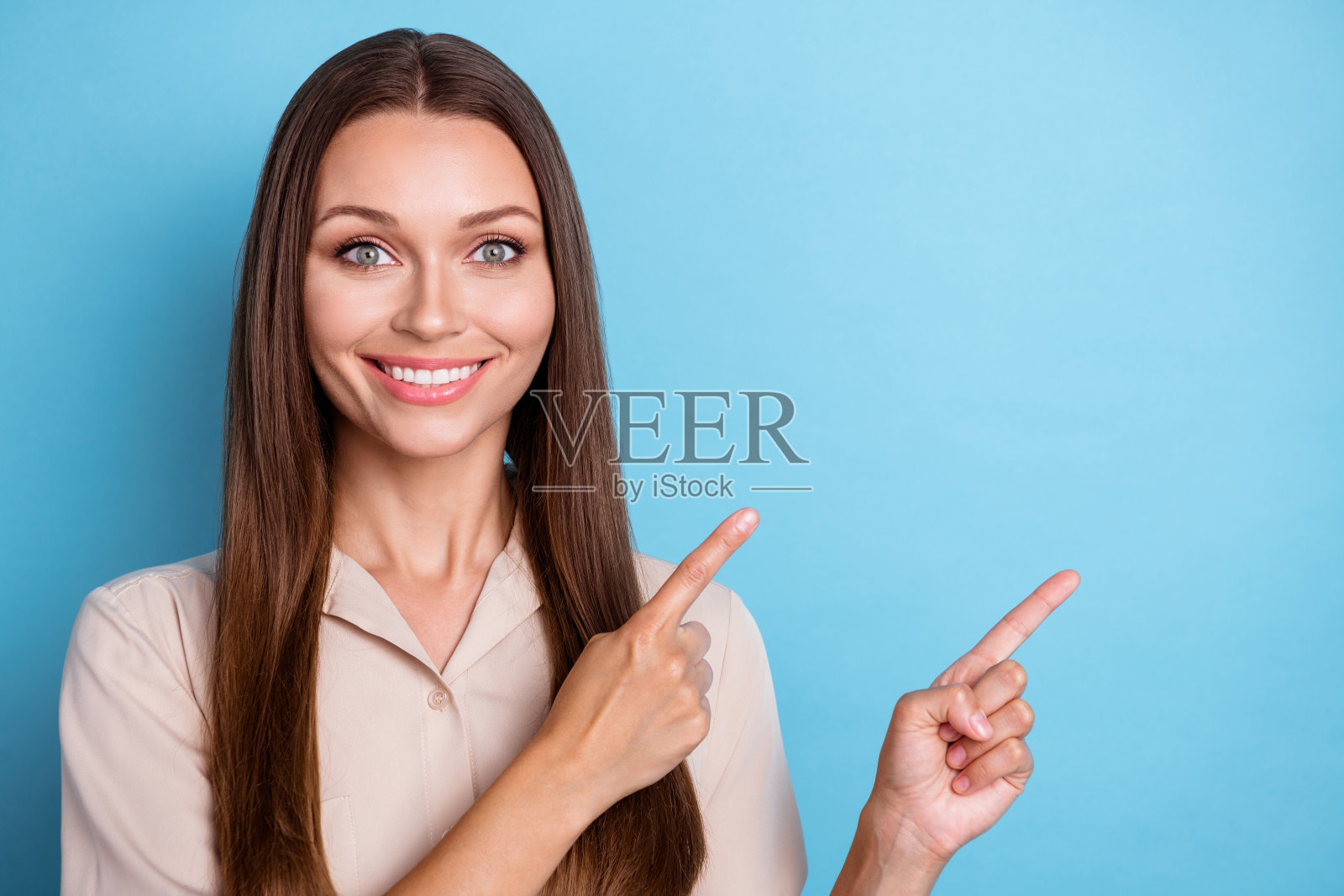 可爱的美丽女孩穿着米色上衣，长发飘逸，面带微笑，指向蓝色背景上的空白处。照片摄影图片