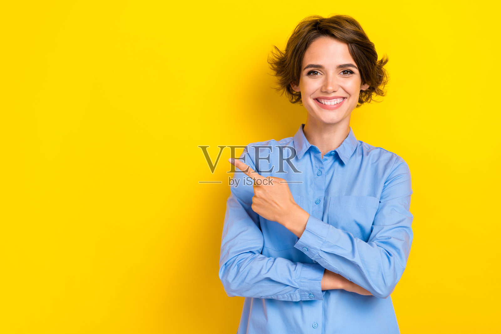 年轻自信女性的肖像照片，穿着蓝色衬衫和正式办公室服装，露出灿烂的微笑，手指指向空白处，背景为黄色。照片摄影图片