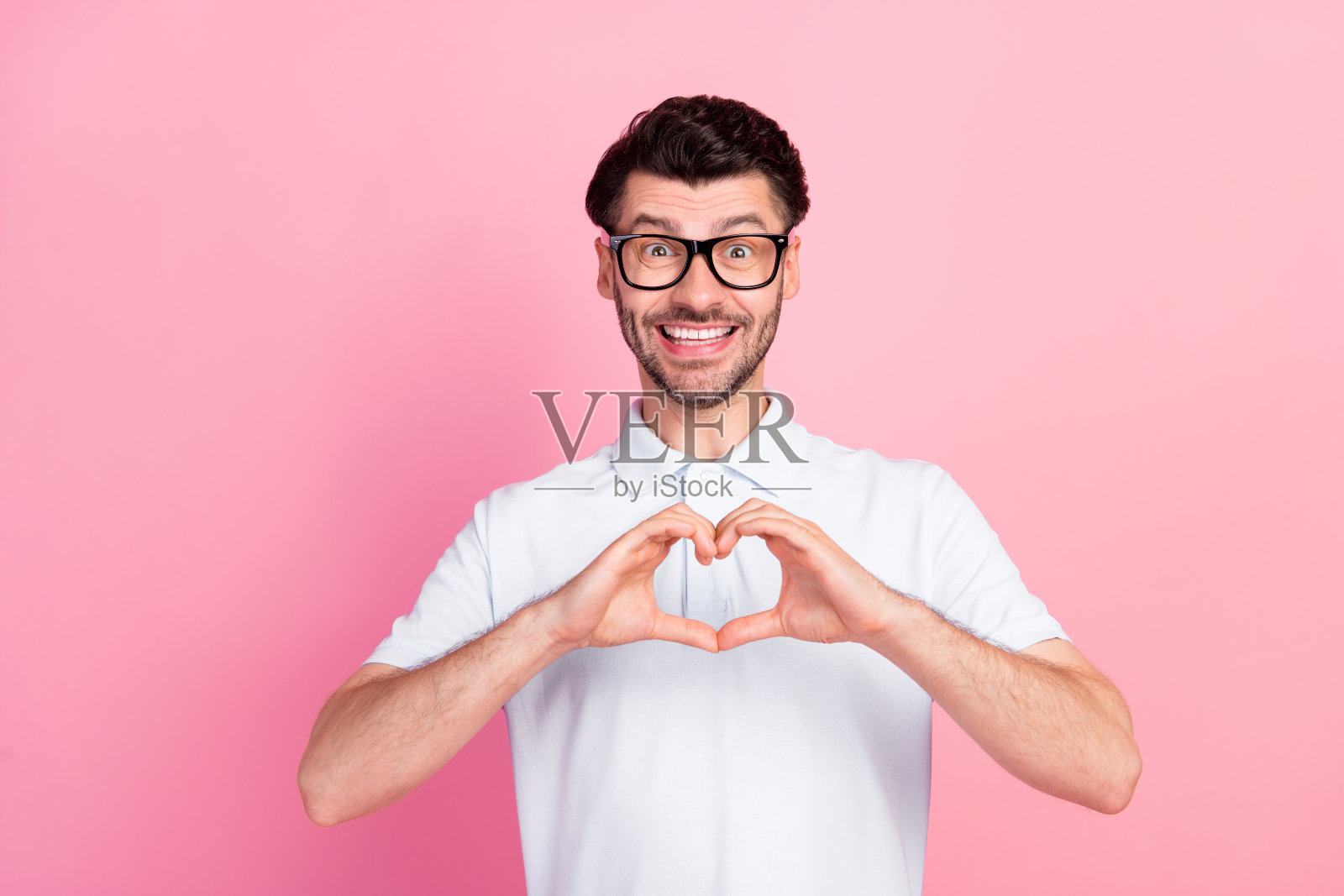 年轻幽默、积极心态的男孩穿着白色T恤，露出灿烂的笑容，做出心形手势，背景为粉色，展现出甜蜜的爱意。照片摄影图片