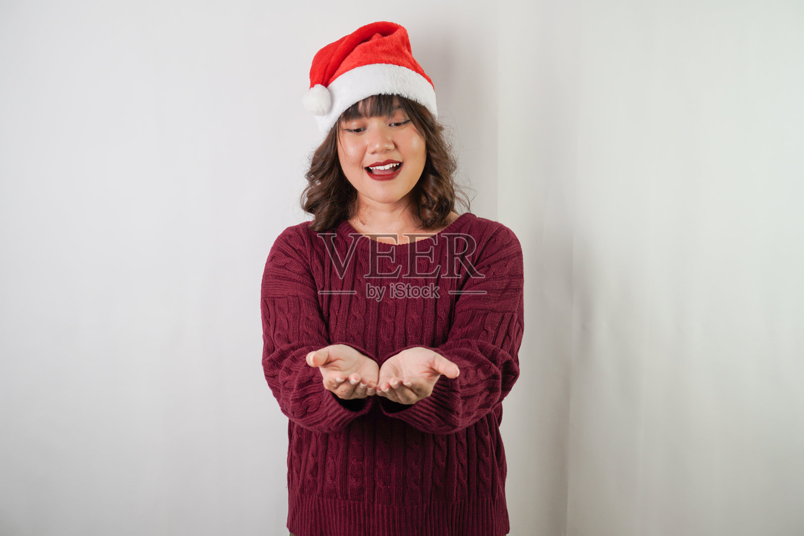 友好的年轻亚洲女性戴着圣诞老人帽，穿着红色长袖毛衣，正展示着空着的手掌，背景为白色。适用于圣诞节和新年派对的概念。照片摄影图片