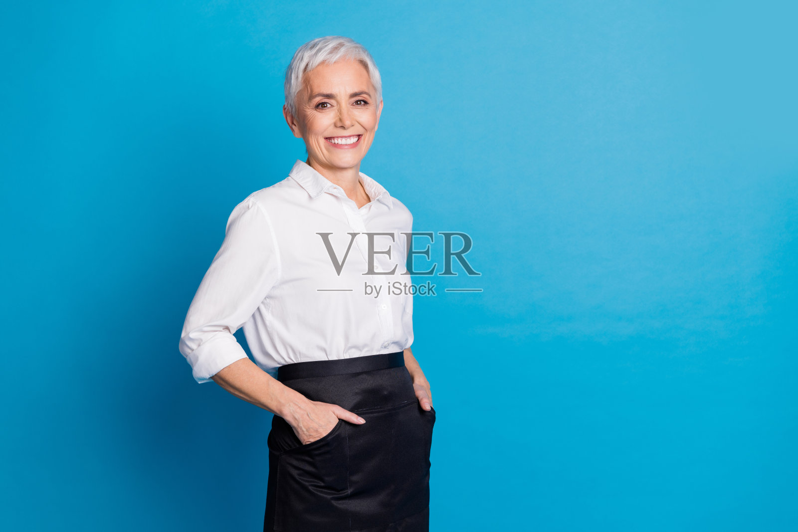 穿着服务员制服的迷人老年女性小咖啡馆老板，面带愉快微笑，背景为蓝色。照片摄影图片