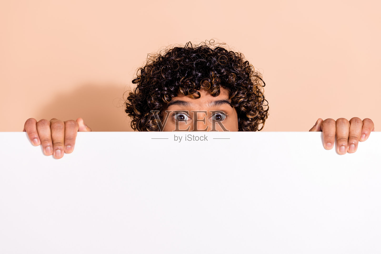 一位墨西哥风格的搞怪害怕的男士，拥有波浪卷发，躲在白色广告牌的空白处，背景为米色。照片摄影图片