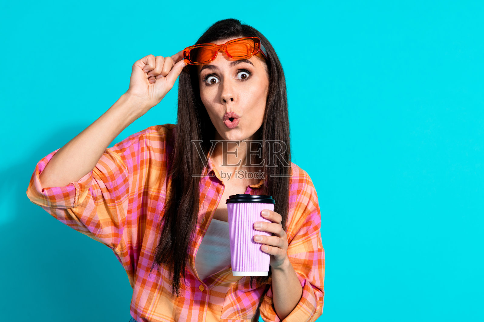 可爱的女士在水绿色背景下，惊讶地摘下太阳镜，手里拿着纸杯，穿着时尚的格子服装。照片摄影图片