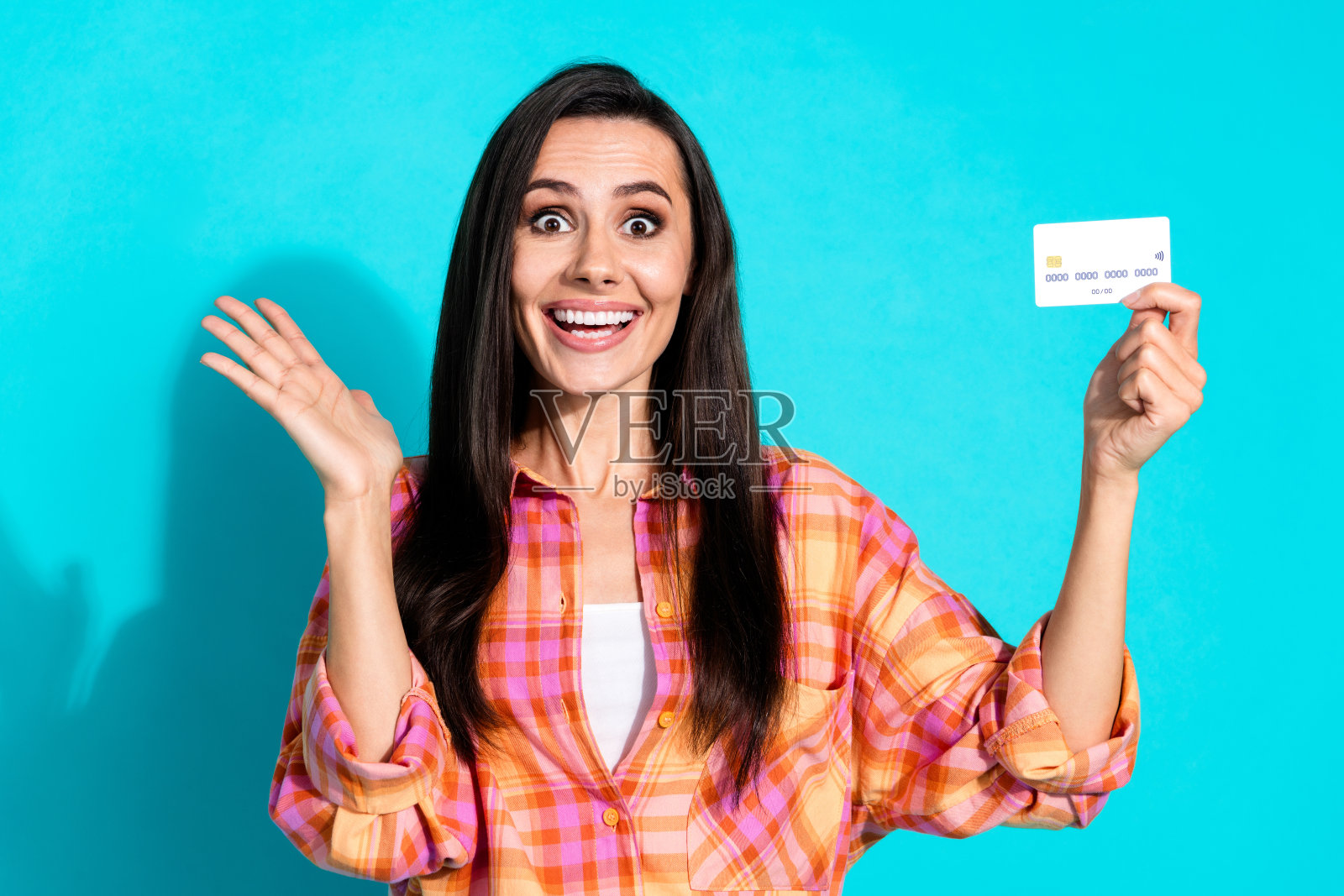 穿着时尚格子衣服的迷人年轻女性，惊讶地拿着信用卡，背景为水绿色。照片摄影图片