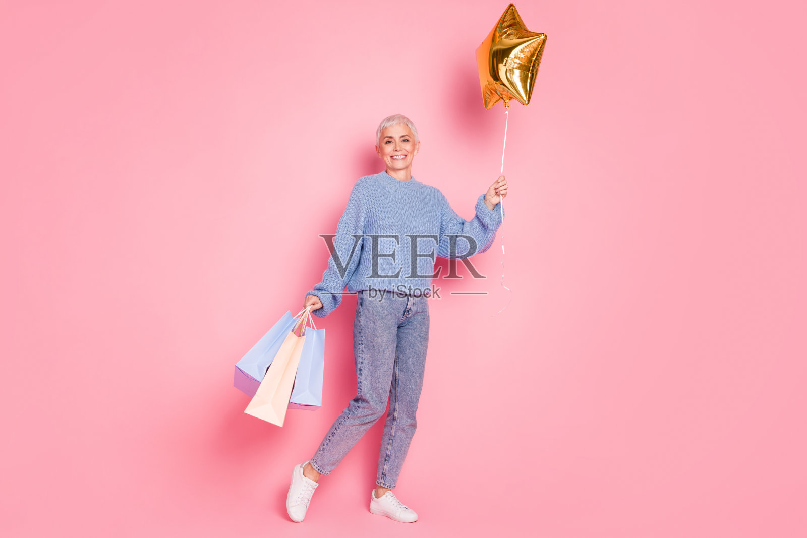 全身照，退休女士舞蹈明星，手持购物袋，穿着时尚的蓝色针织服装，背景为粉色。照片摄影图片