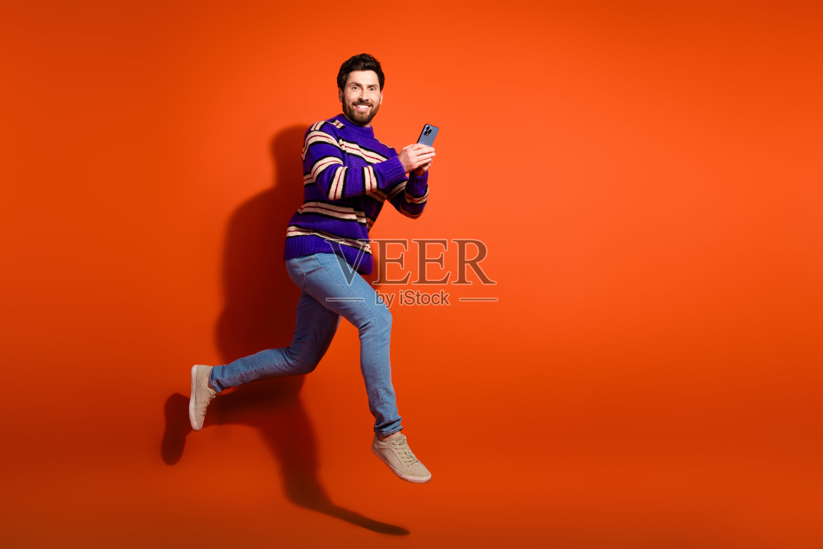 全身照，吸引人的年轻男子穿着时尚的紫色条纹服装，手持小工具，正在奔跑，背景为橙色。照片摄影图片