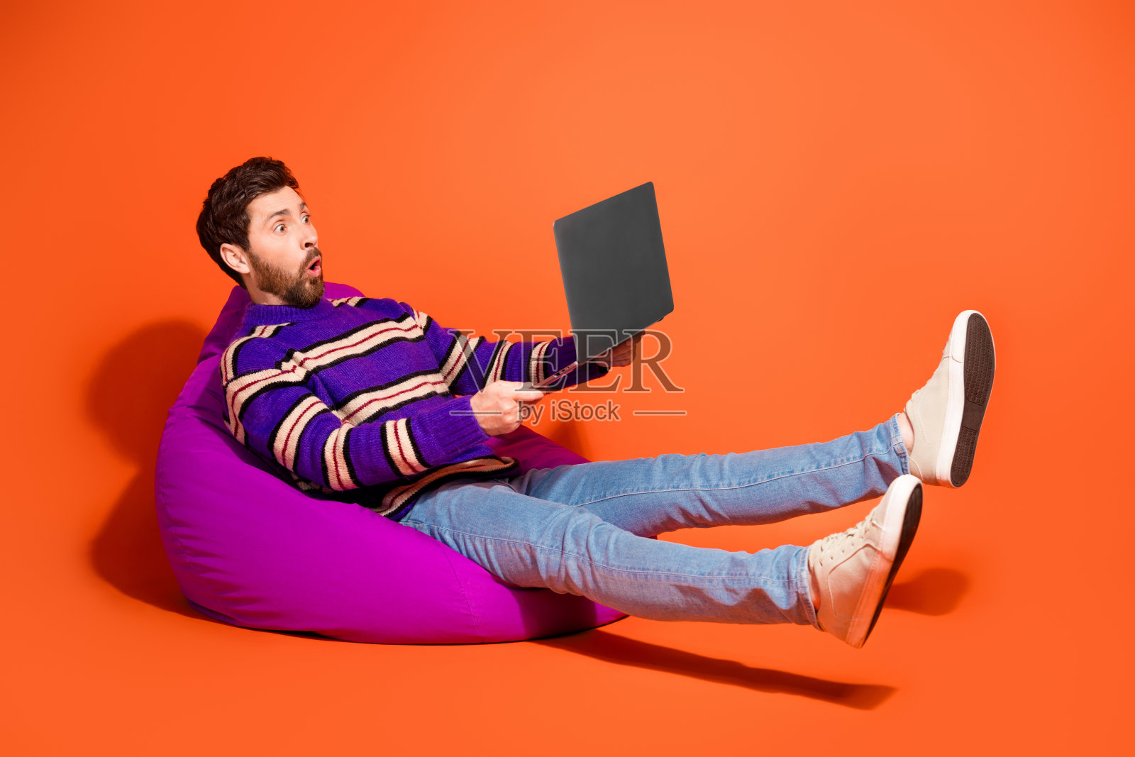 全身照，帅气的年轻男孩坐在豆袋椅上，惊讶地拿着笔记本电脑，穿着紫色条纹衣服，背景为橙色。照片摄影图片
