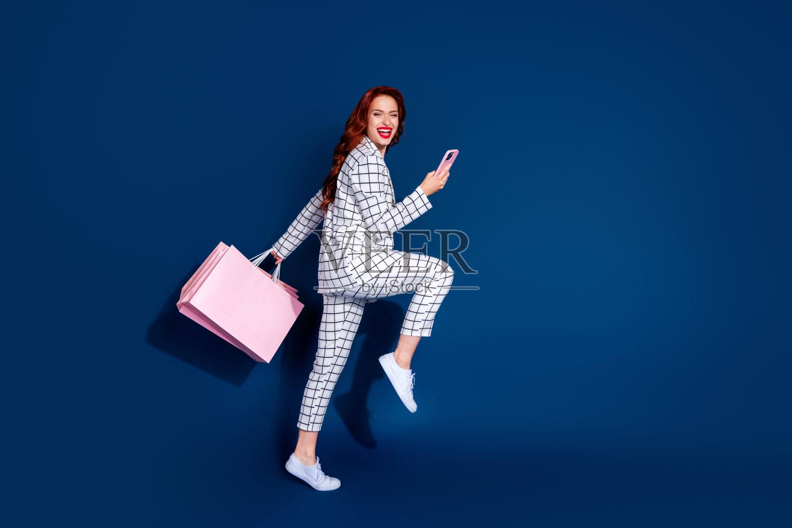 年轻女子，红色头发，穿着时尚的格子西装，愉快地展示她的购物战利品，同时手持智能手机。照片摄影图片