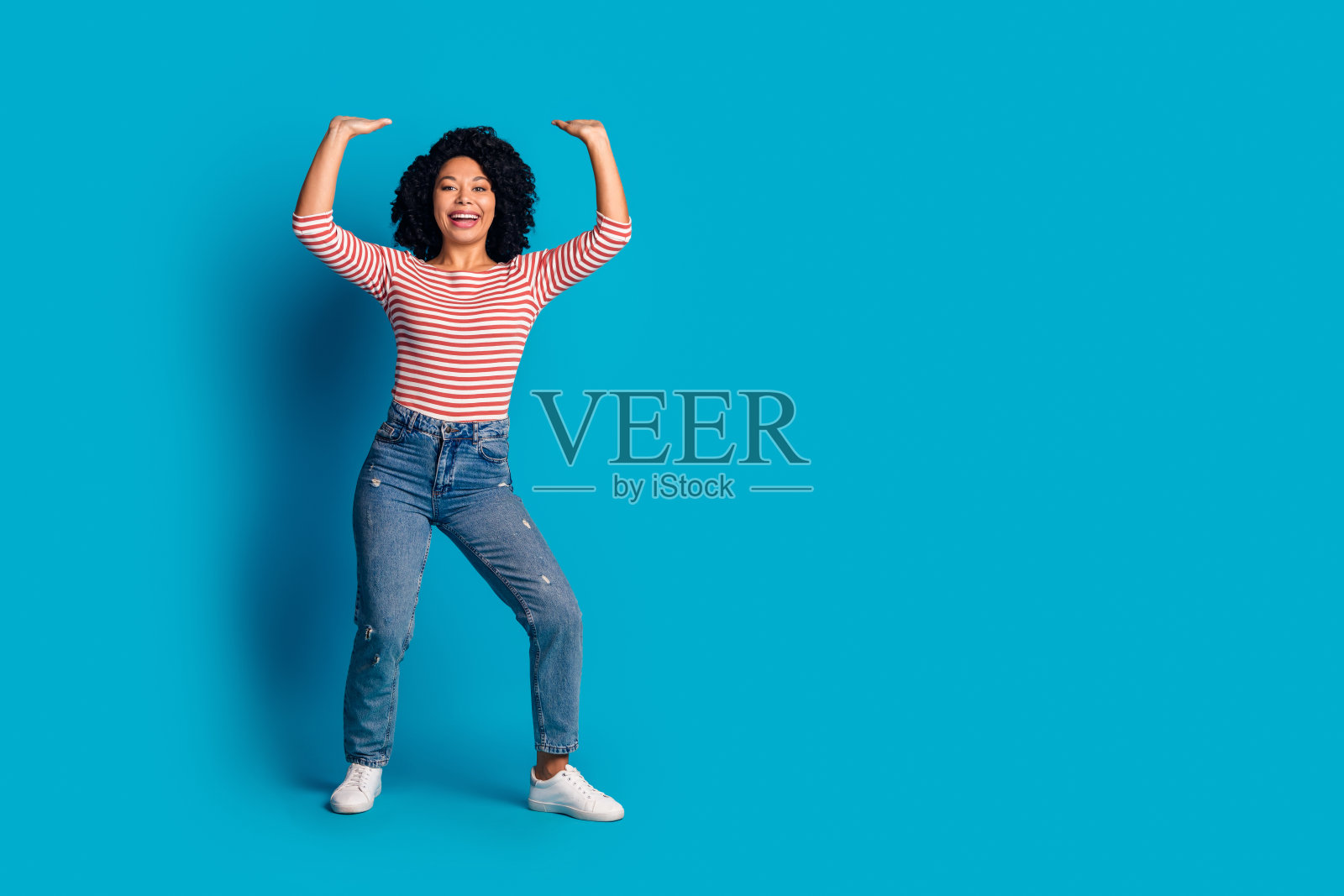 可爱的幽默乐观女孩穿着时尚衣服，手举过头，手中空无一物，背景为蓝色。照片摄影图片