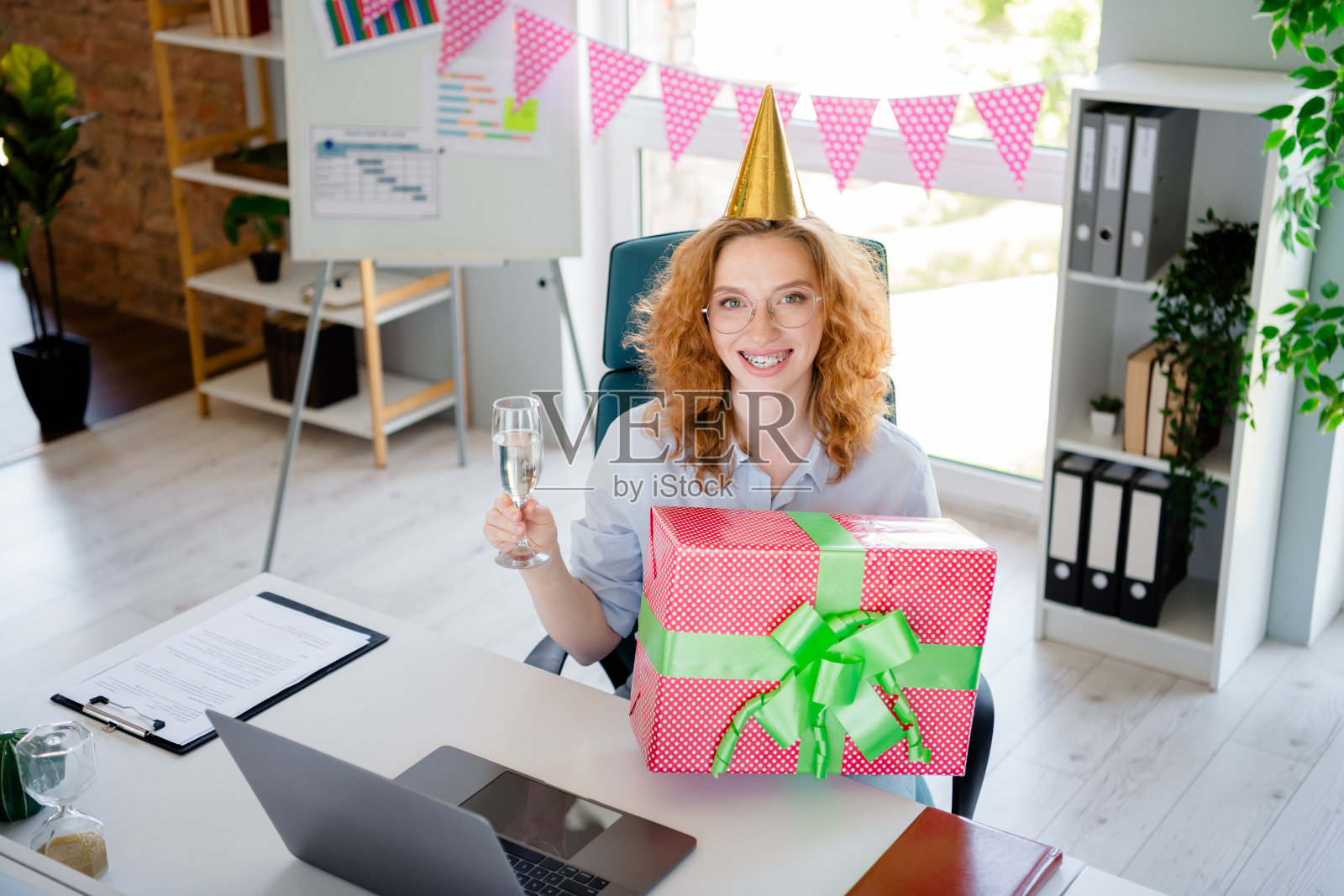 年轻女士在舒适的初创公司办公室内庆祝生日，手持香槟，身穿正式商务服装，旁边有礼品盒。照片摄影图片