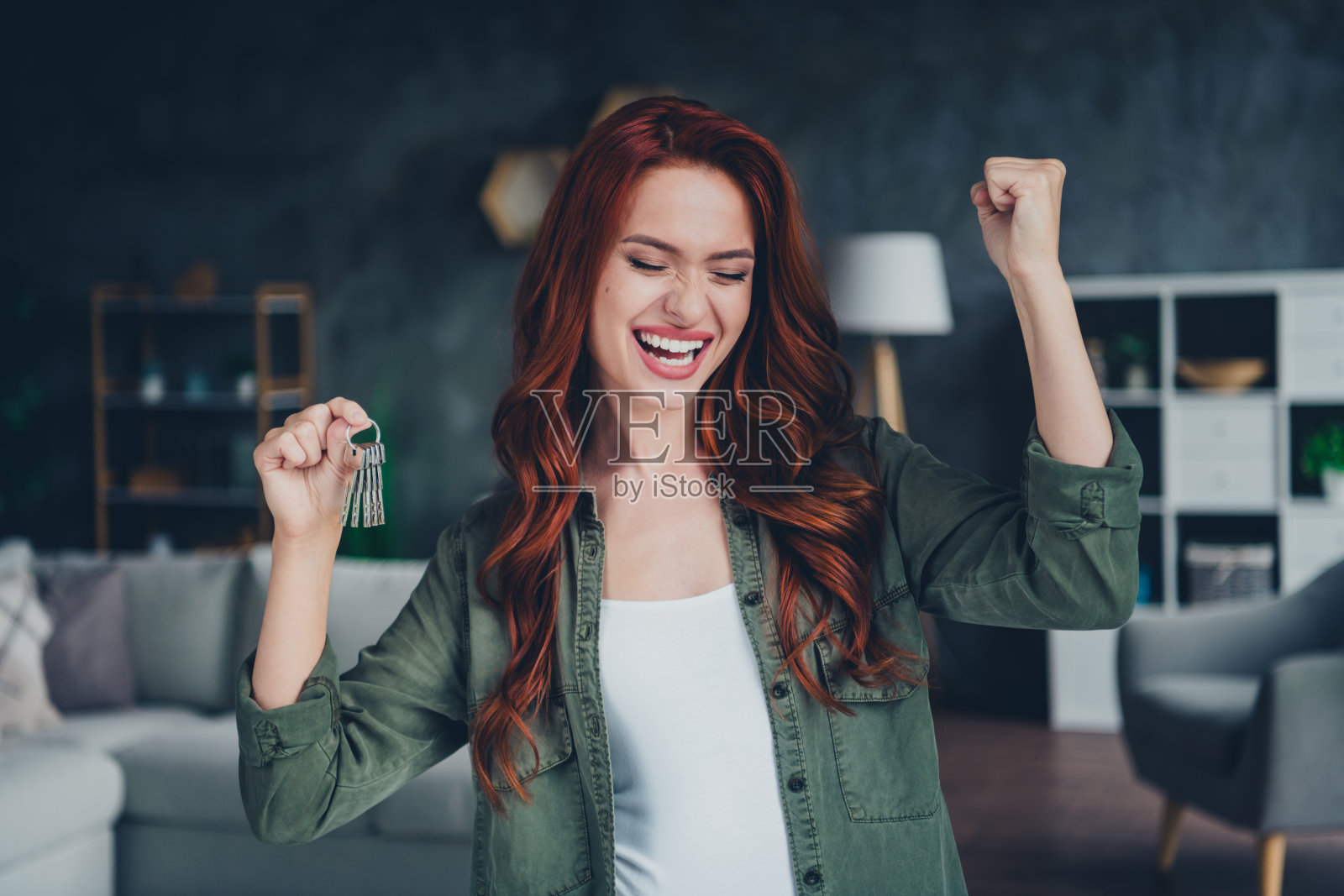 年轻的红发女孩在现代舒适的公寓室内举起拳头，穿着卡其色衣服，显得很开心。照片摄影图片